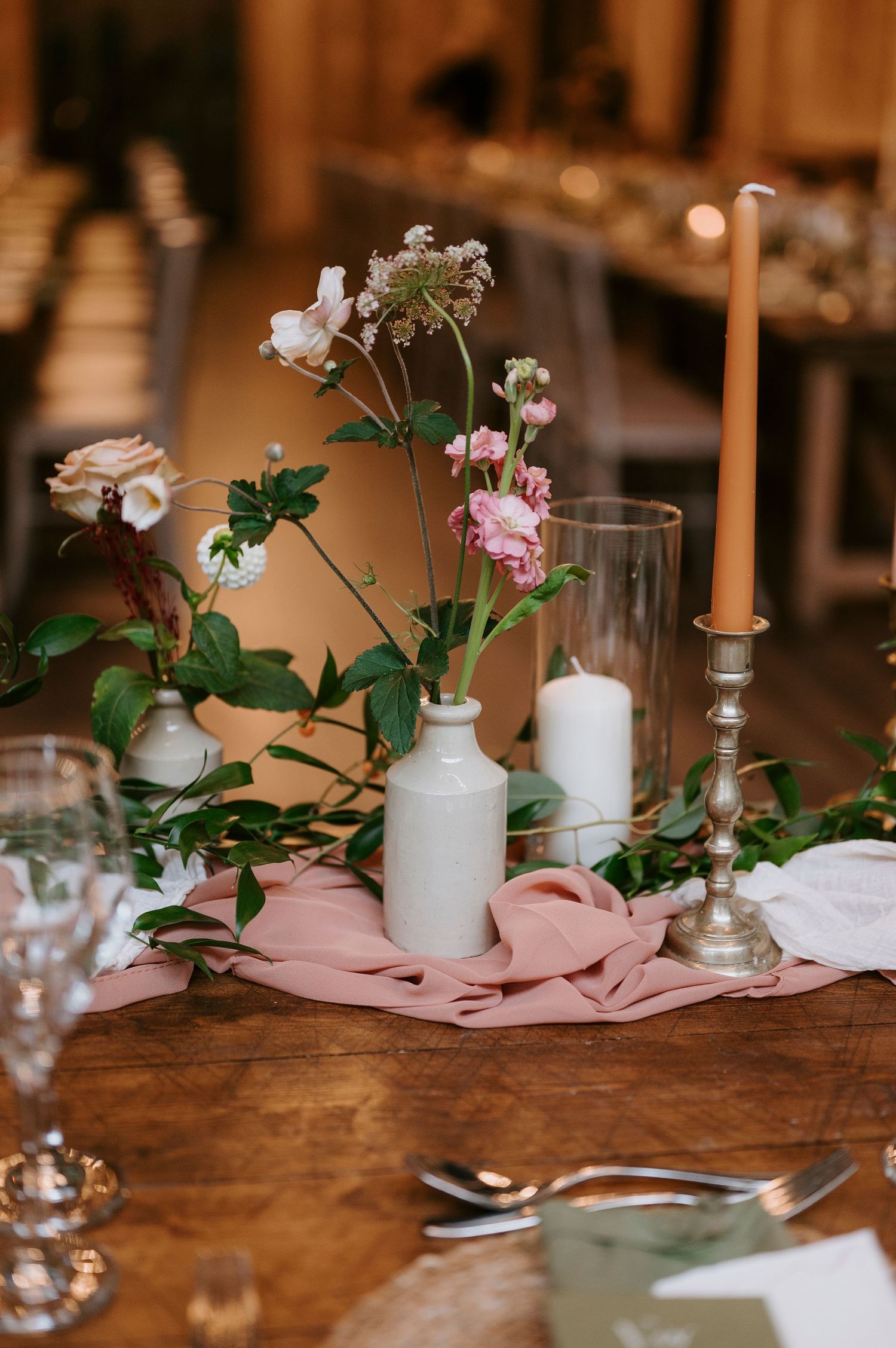 Elegant table setting with white candles and pink floral arrangements on rustic wooden surface for wedding reception.