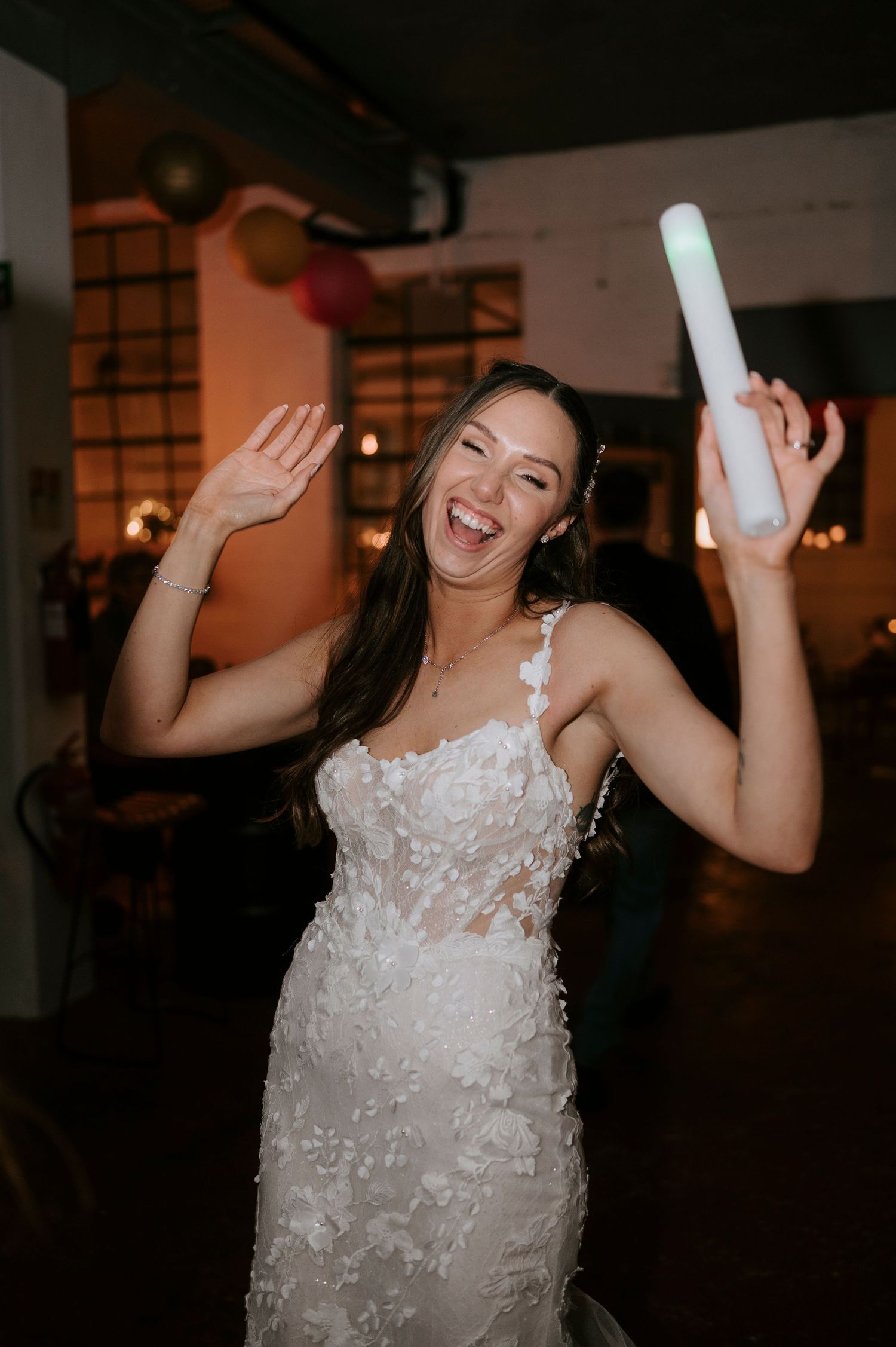 A joyful bride in a floral lace gown dances with a glow stick at her wedding reception.