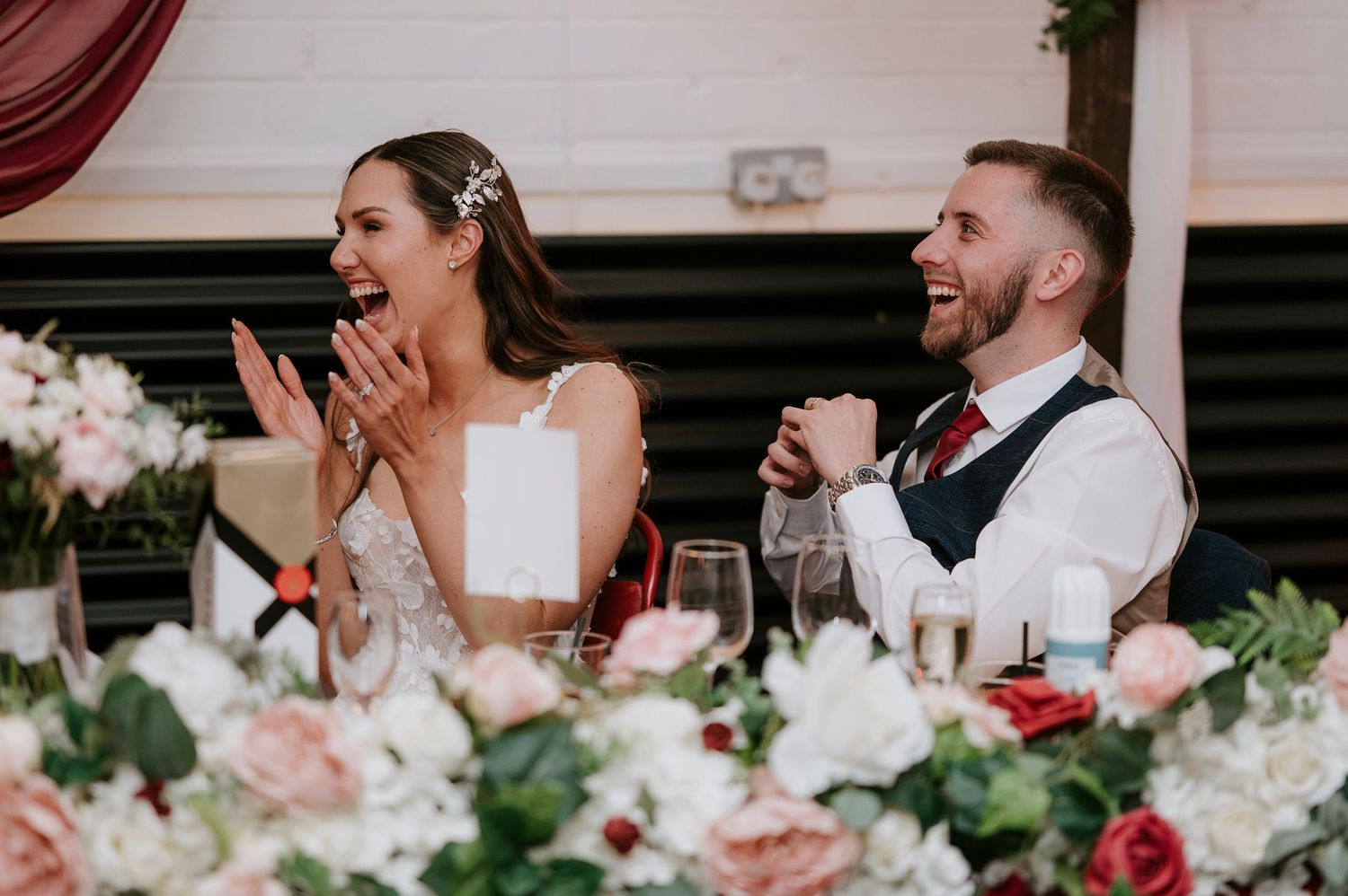 Bride and groom laughing joyfully at their wedding reception surrounded by pink and red floral arrangements.