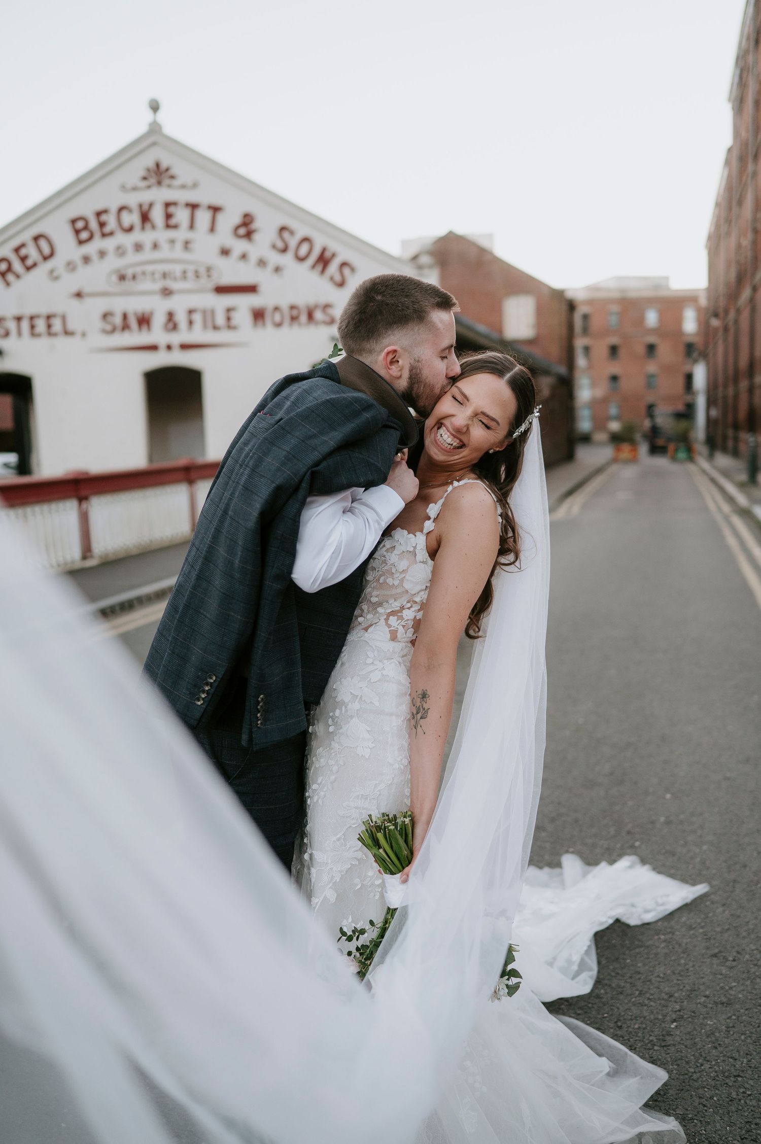 Bride and groom share a joyful kiss on a cobblestone street outside Fred Beckett & Sons Steel Works.