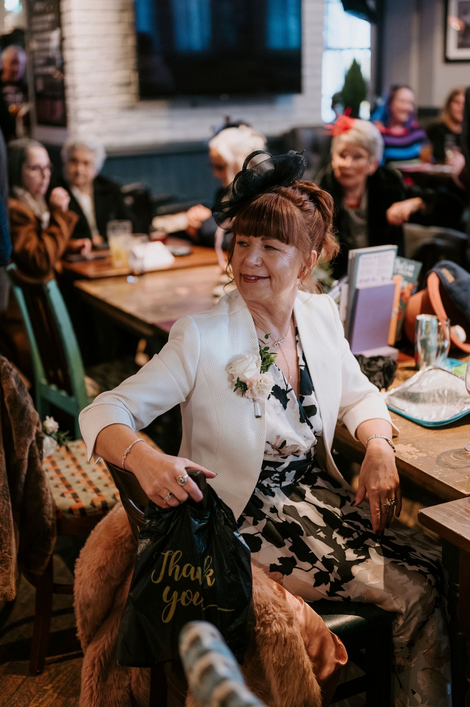 Woman in white blazer smiling at wedding reception, holding a Thank You gift bag, with guests seated at tables behind her.