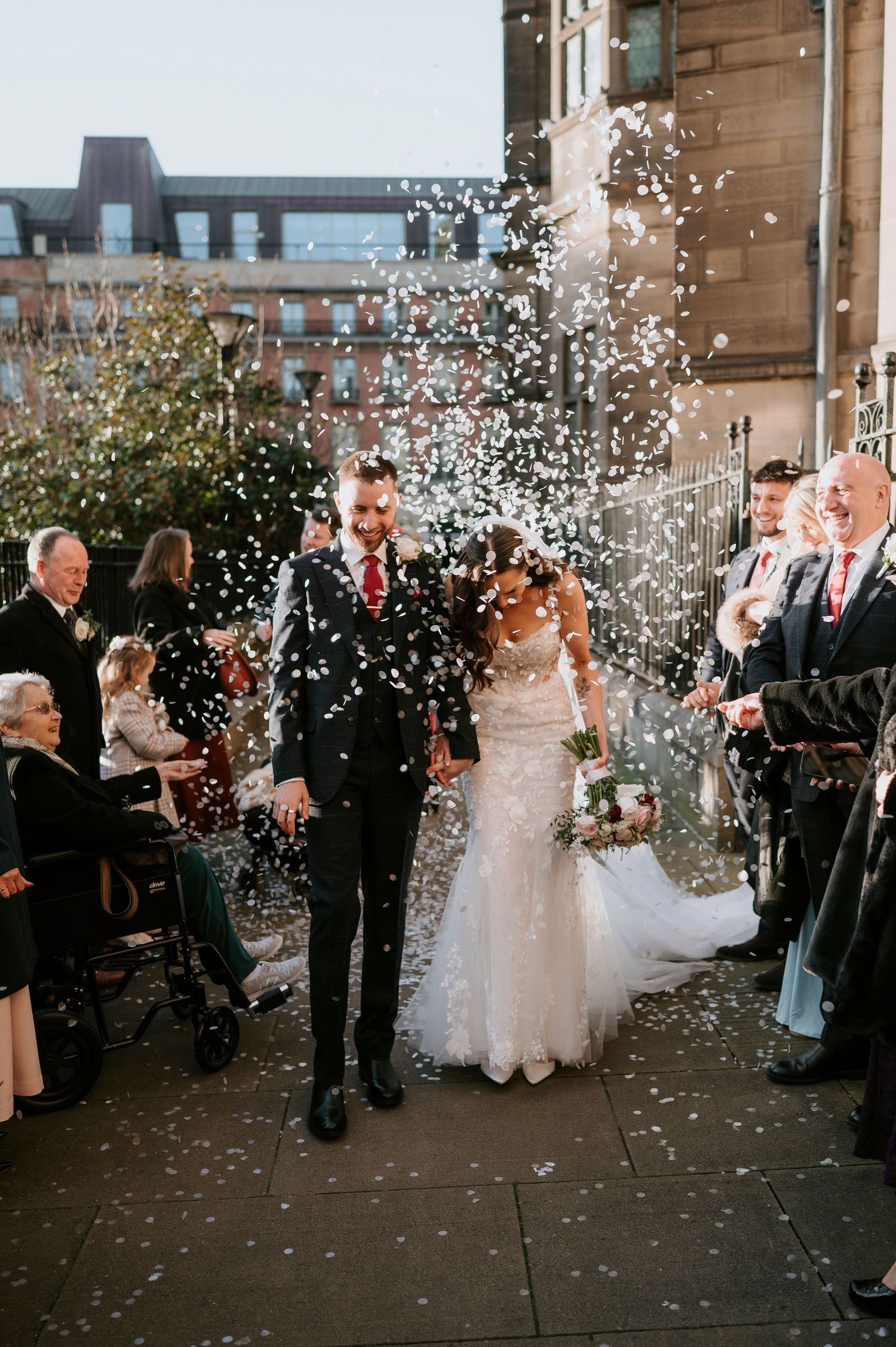 Newlywed couple walks through confetti shower surrounded by cheering guests outside a brick building.