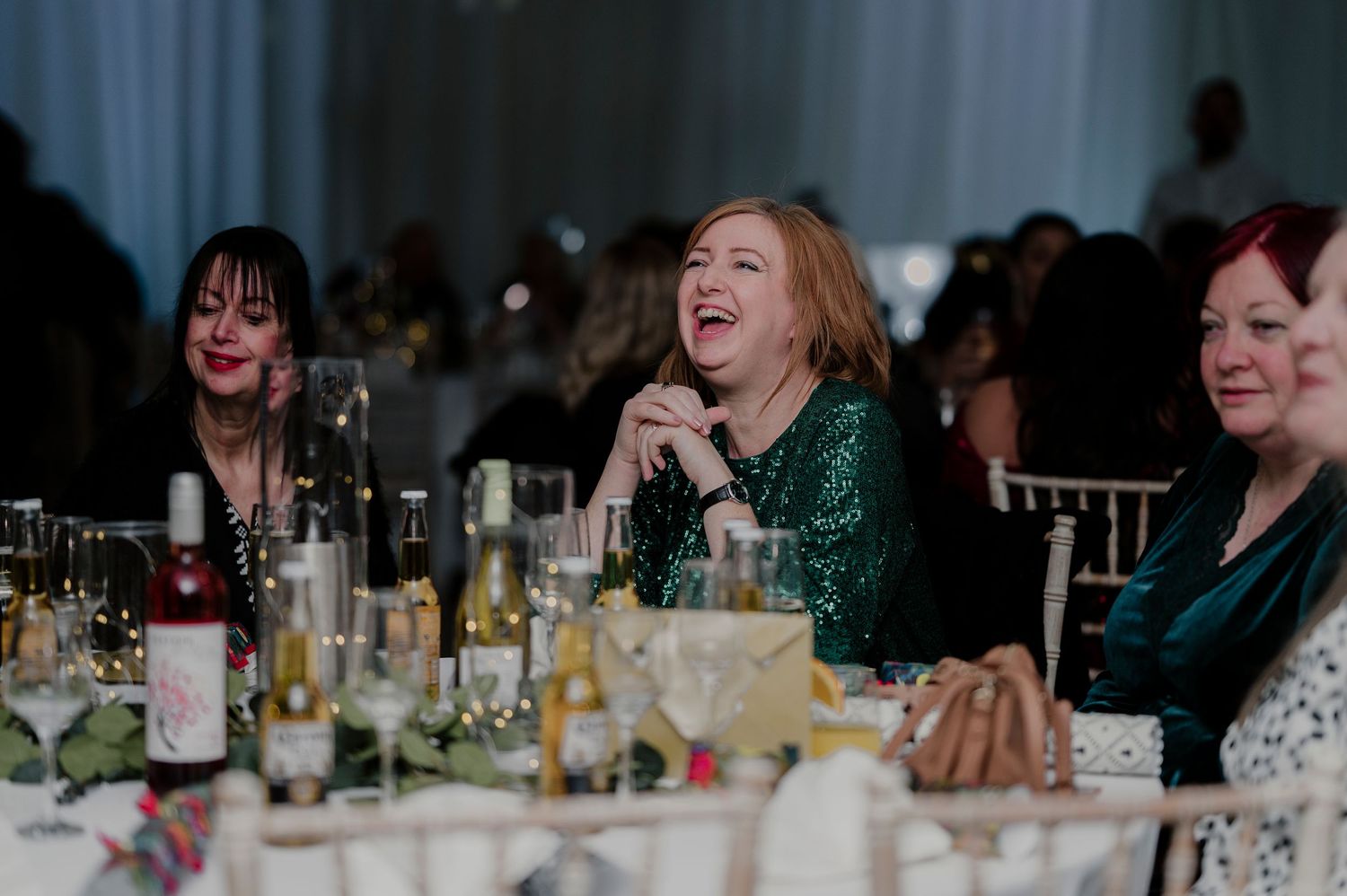 Wedding guests laugh and socialize around a table filled with drinks and glassware at a reception celebration.