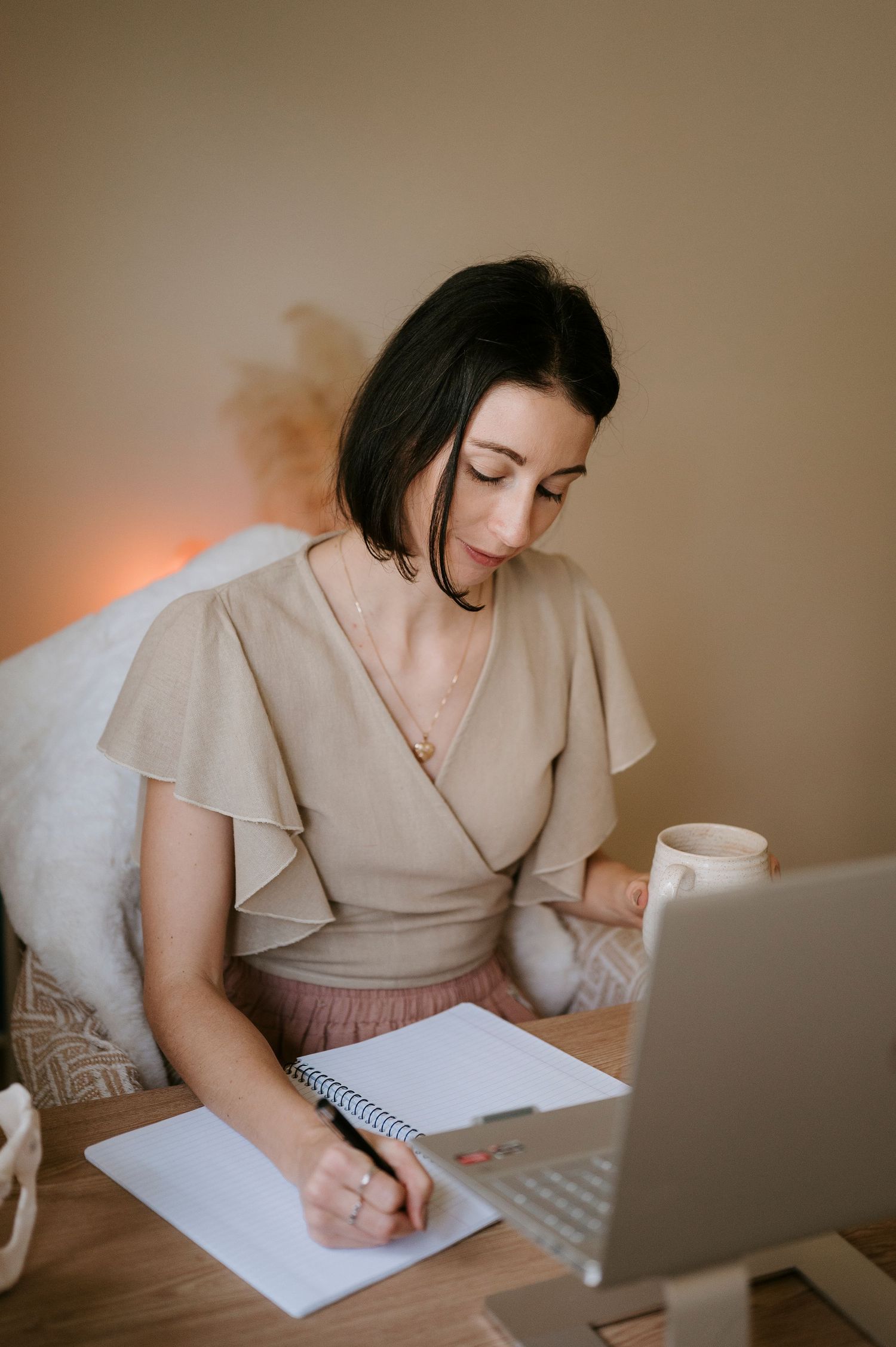 Person in beige top writes in notebook while working at desk with laptop and coffee cup in warm-lit room.