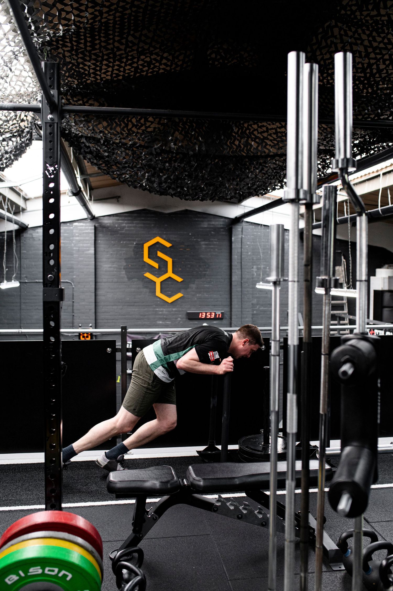 Athlete performs angled push-up on bench in modern gym with orange SC logo on dark wall and weight plates visible.