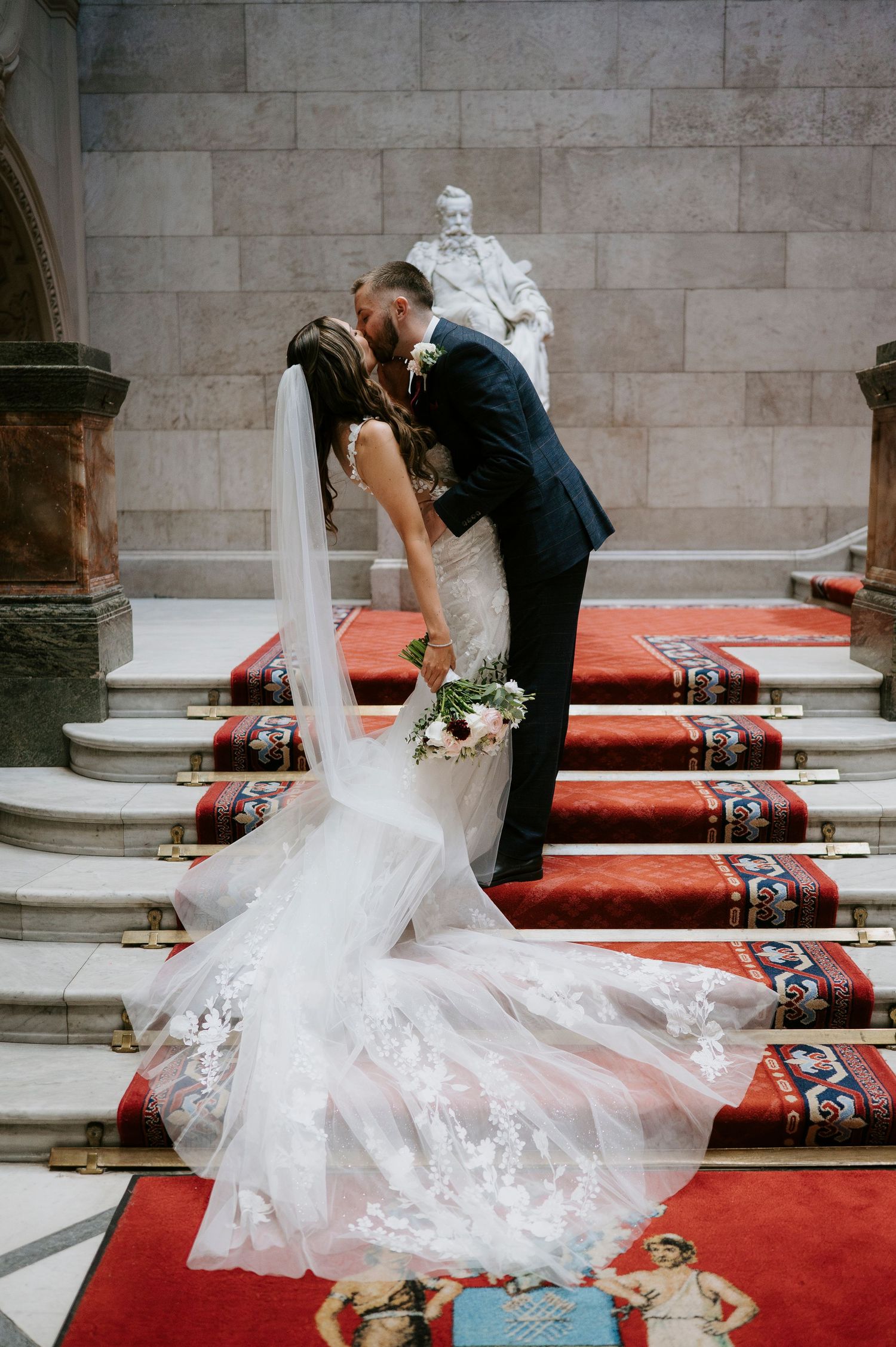 Bride and groom share a romantic kiss on red-carpeted stairs in an elegant venue, with lace wedding dress train spread beautifully.
