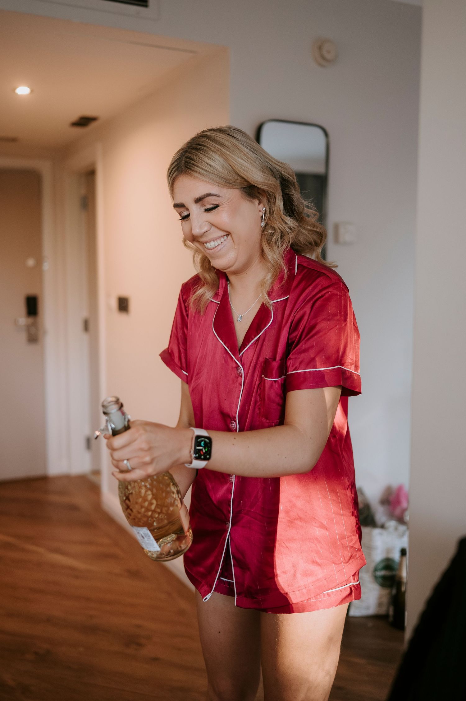 Smiling blonde woman in red satin pajamas opening a champagne bottle in a bright hotel room.