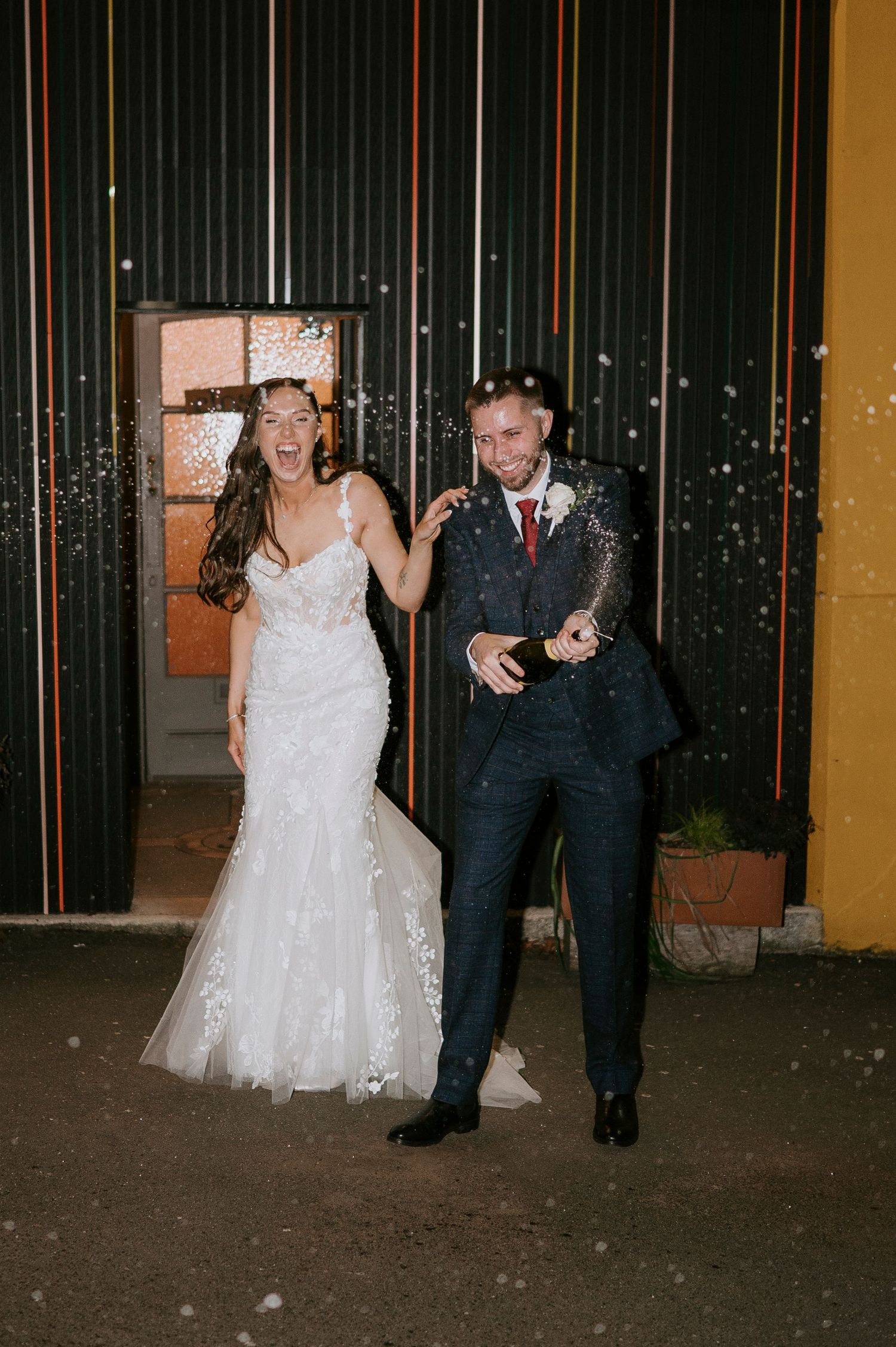 Joyful bride and groom celebrate with champagne confetti outside their wedding venue at night.