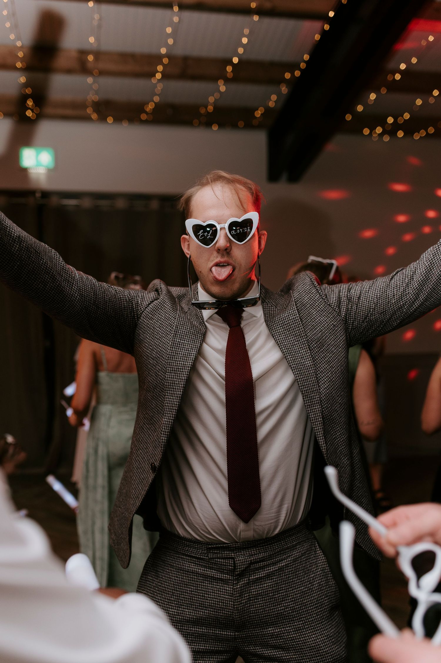 Person in grey suit and red tie dancing energetically at a party with heart-shaped sunglasses and disco lights.