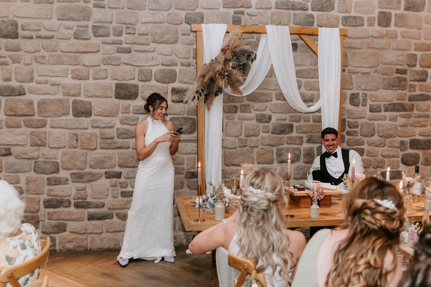 A wedding reception in a rustic stone-walled venue with guests seated at long tables decorated with floral centerpieces.