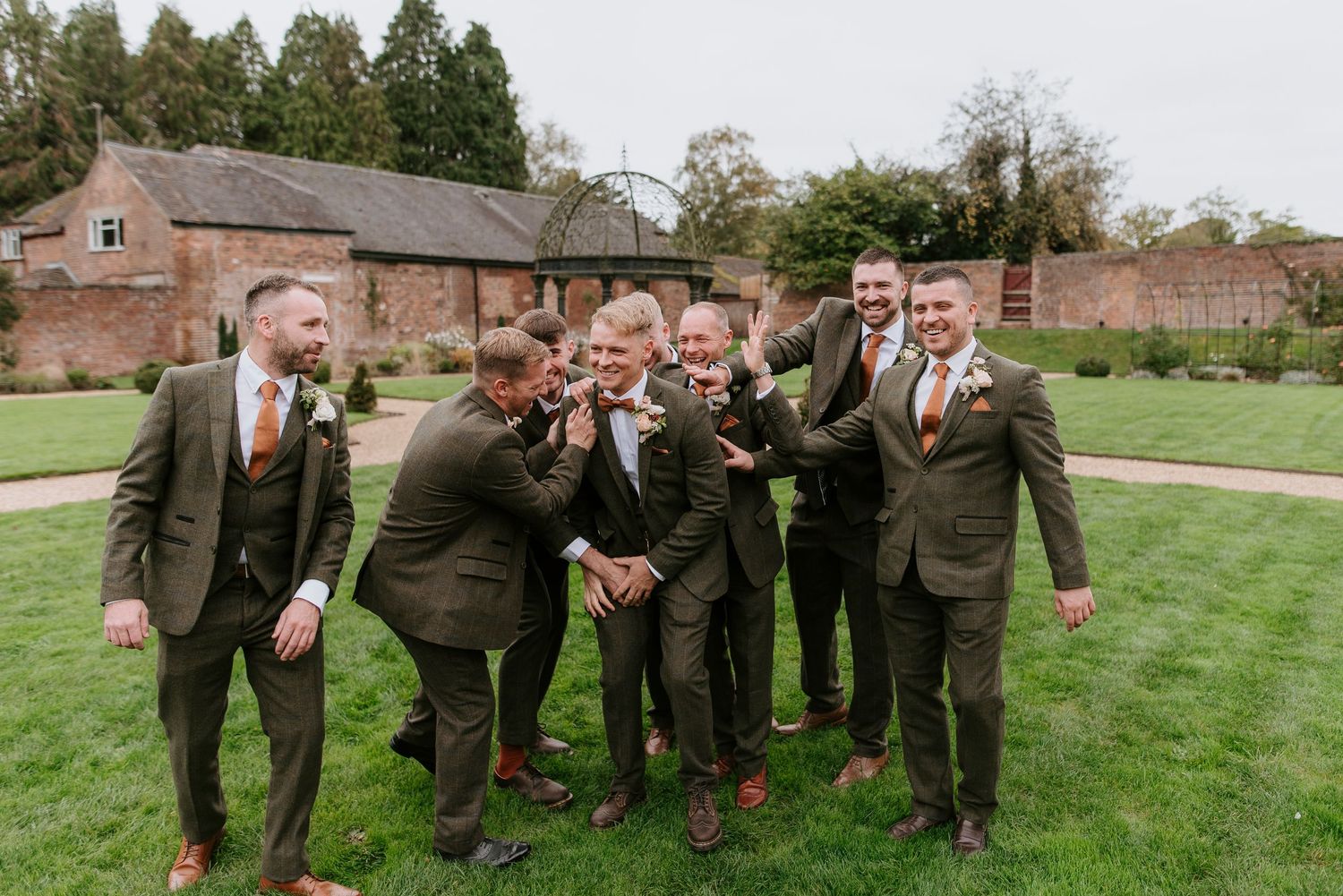 A group in matching olive green suits laughing together on a lawn in front of rustic brick buildings.
