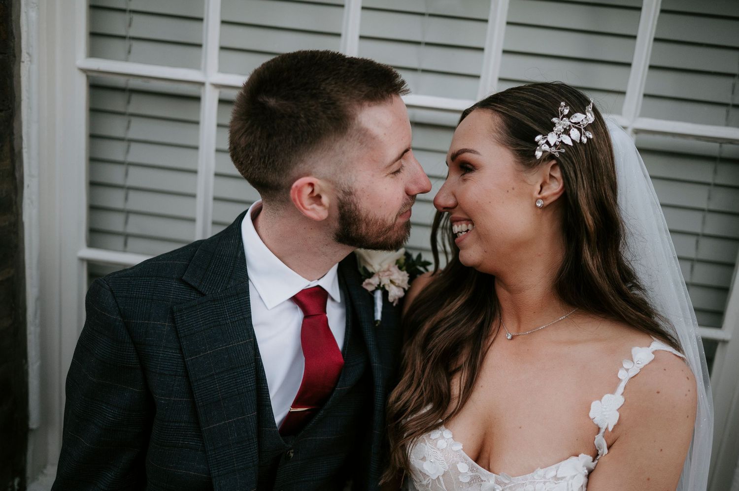 Bride and groom share an intimate moment, smiling and almost kissing, bride wearing floral hair accessory.