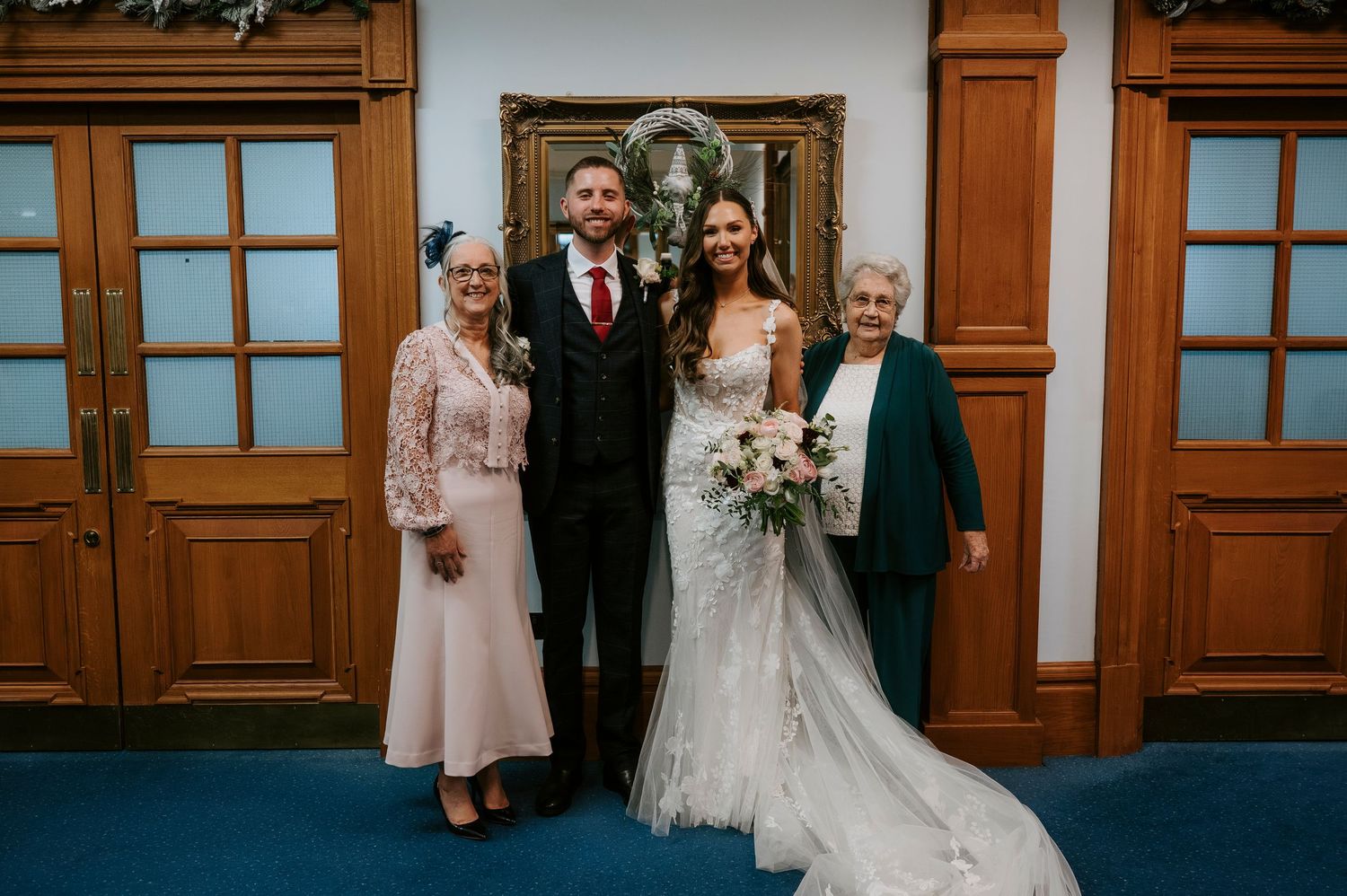 Bride and groom pose with two elderly women in elegant attire at a formal wedding ceremony indoors.