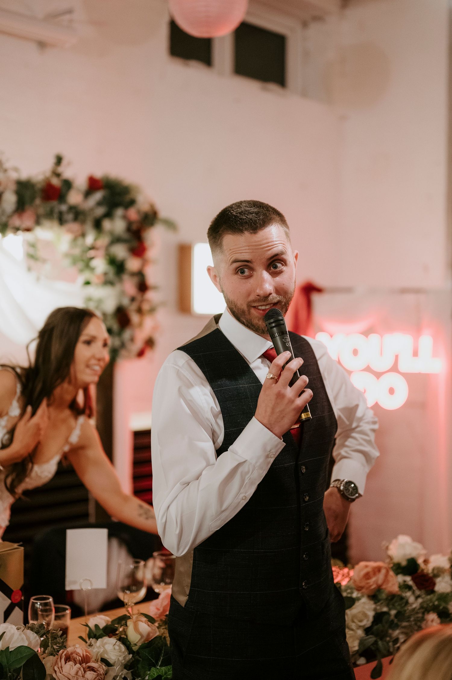 Groom in white shirt and dark vest delivers speech into microphone at wedding reception with neon sign backdrop.