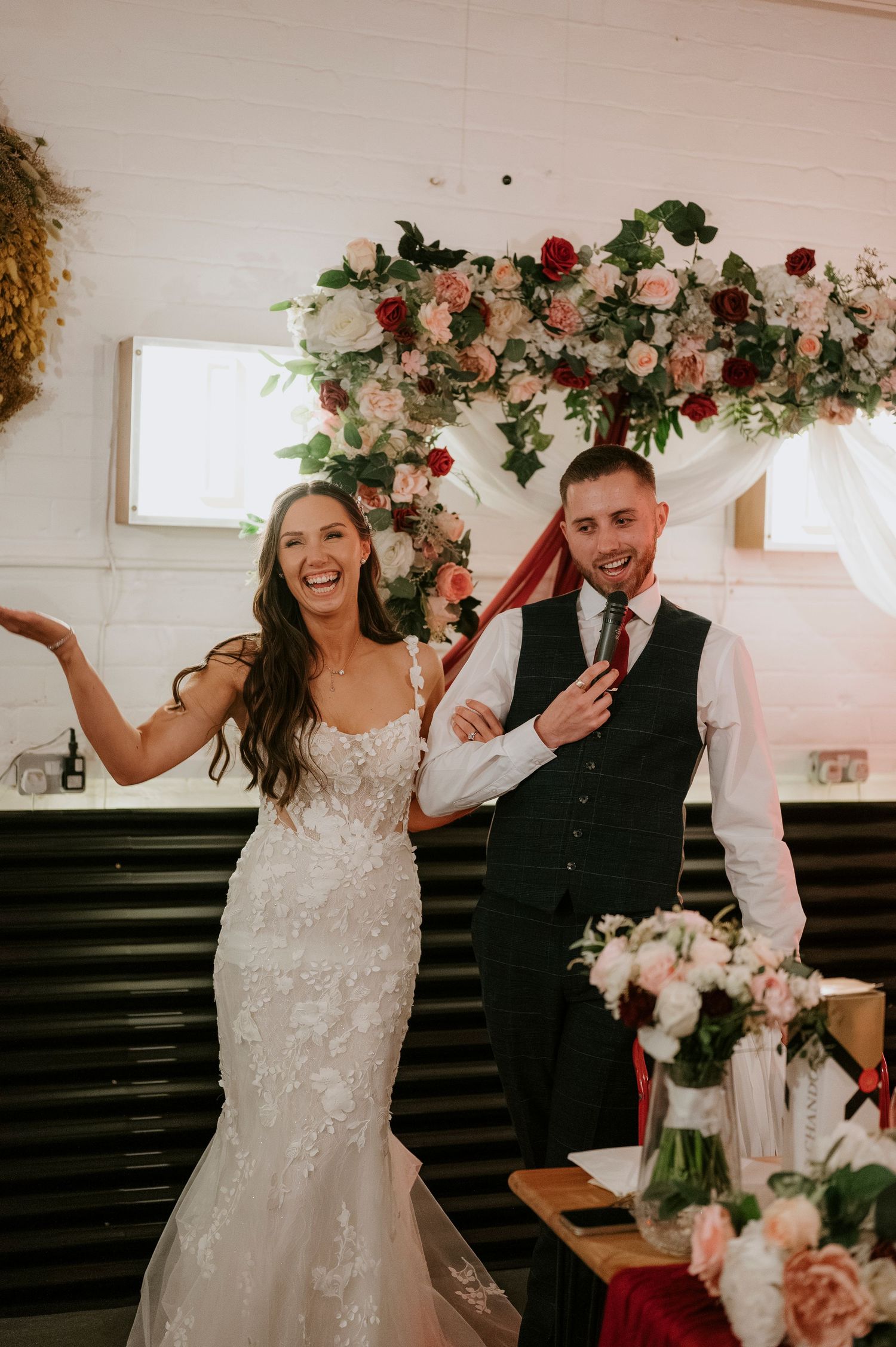 Joyful bride in lace gown and groom in dark vest celebrate at their wedding reception with floral arch backdrop.