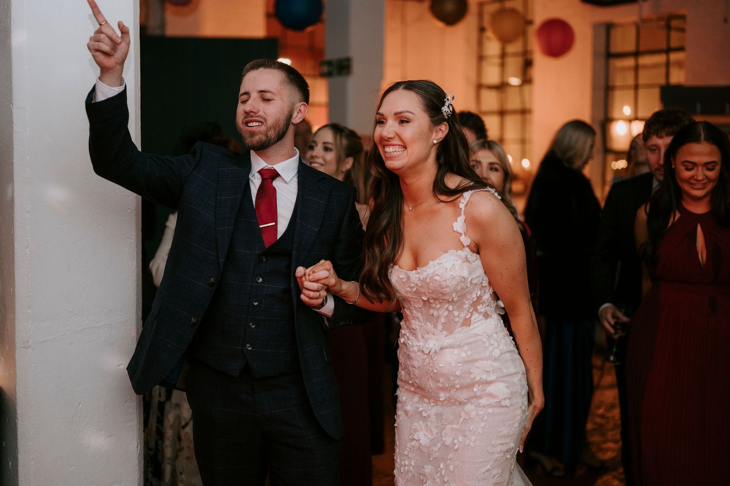 Smiling bride in lace gown enters wedding reception holding grooms hand as guests celebrate.