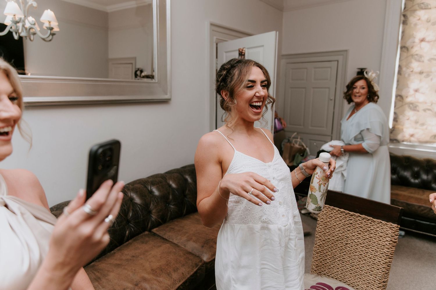 A joyful moment captured in a bridal suite with a leather couch and mirror as someone tries on a white dress.