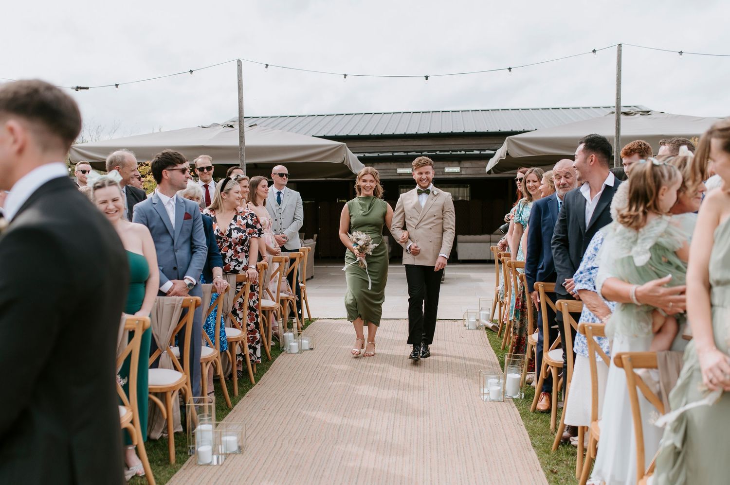 A wedding ceremony processional on an outdoor deck with guests seated in wooden chairs along the aisle.