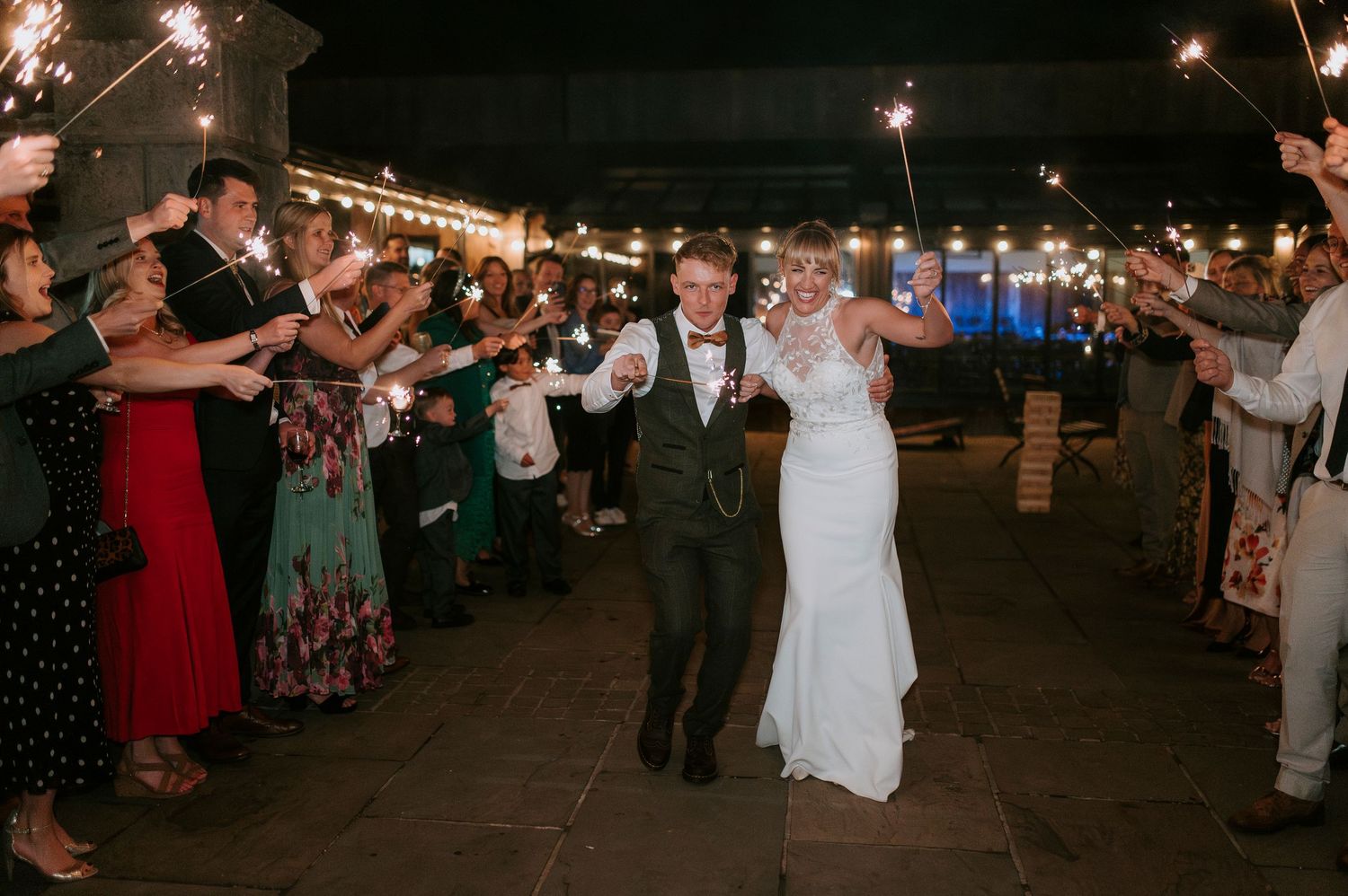 Wedding guests hold sparklers creating a glowing tunnel for the newlyweds during an outdoor nighttime celebration.