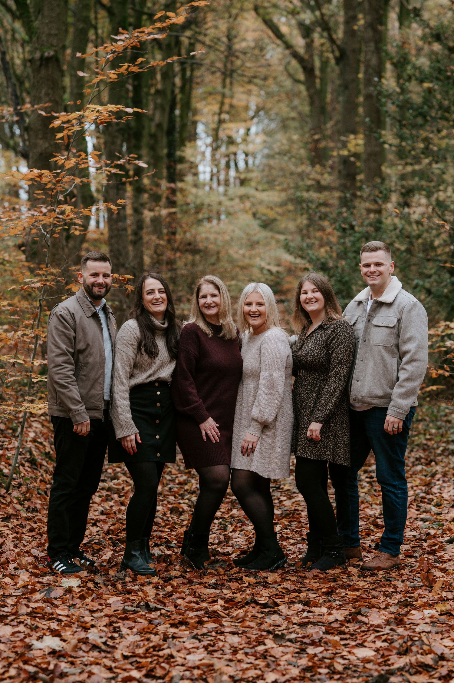 Family group poses together on fallen autumn leaves in a forest setting wearing coordinated neutral tones.