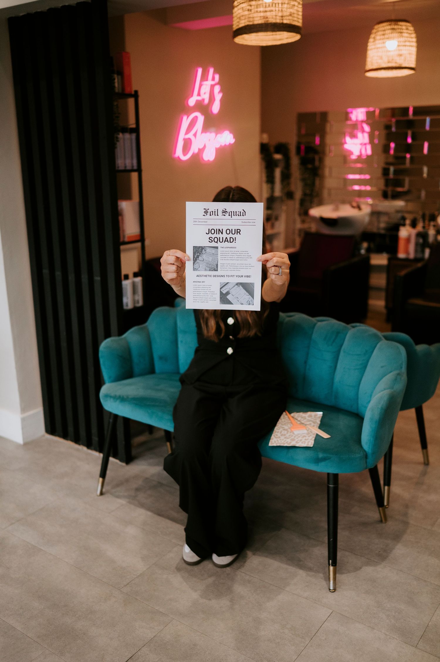 Someone holding a paper document while sitting in a teal velvet chair with pink neon signs glowing in the background.