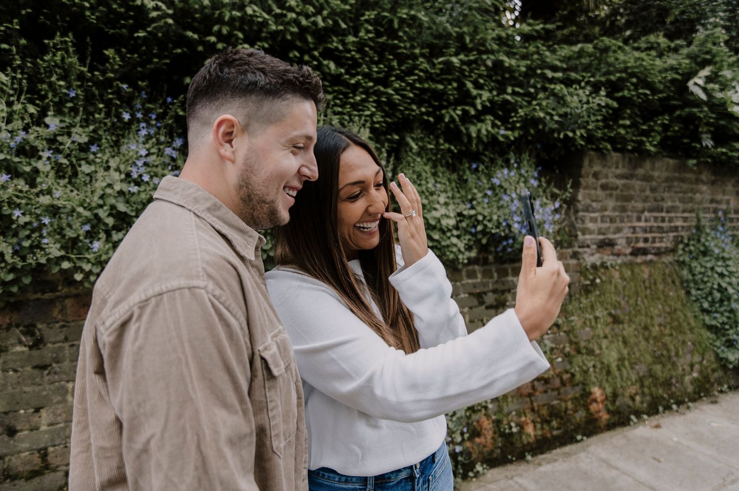 A couple shares a joyful moment while taking a selfie together in front of an ivy-covered wall.