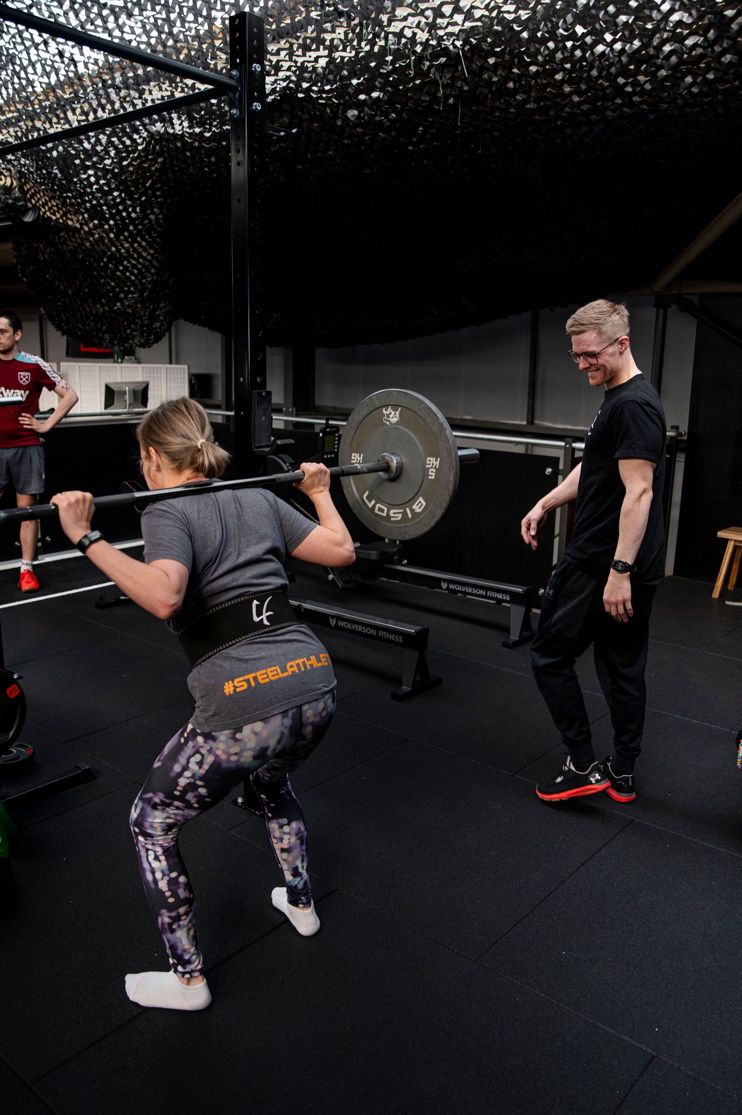 A trainer supervises a person performing barbell squats during a workout session in a dark gym setting.