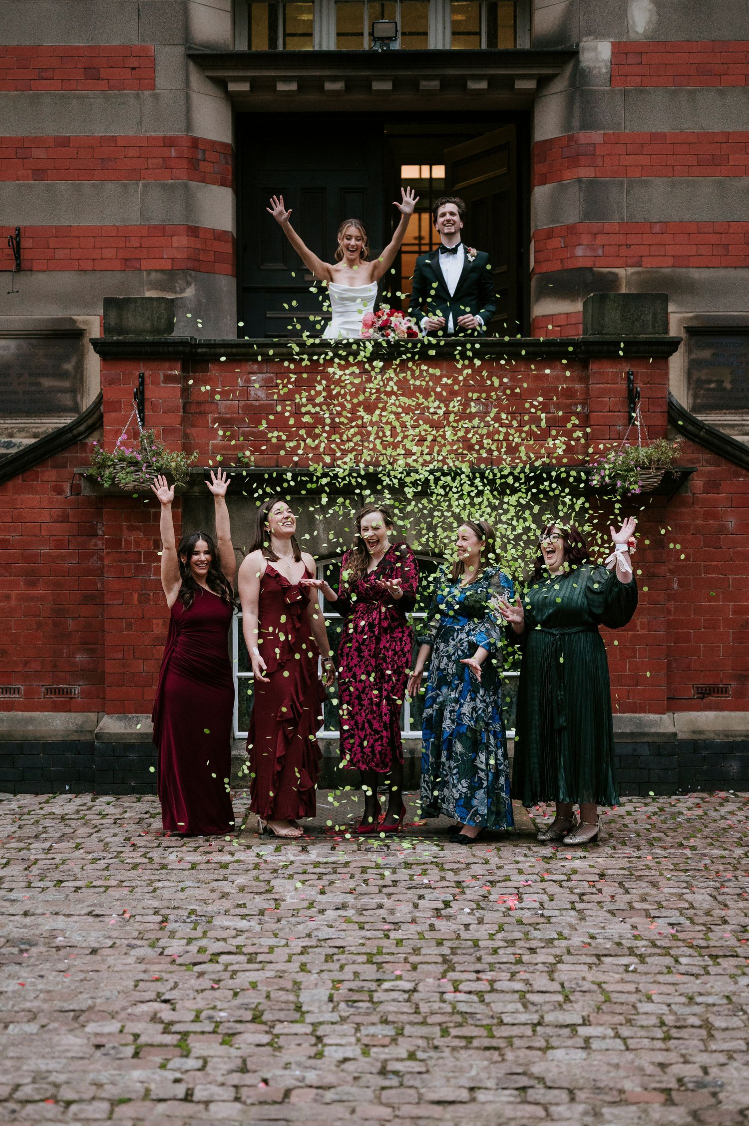 Wedding party stands in front of brick building as confetti rains down in celebration.