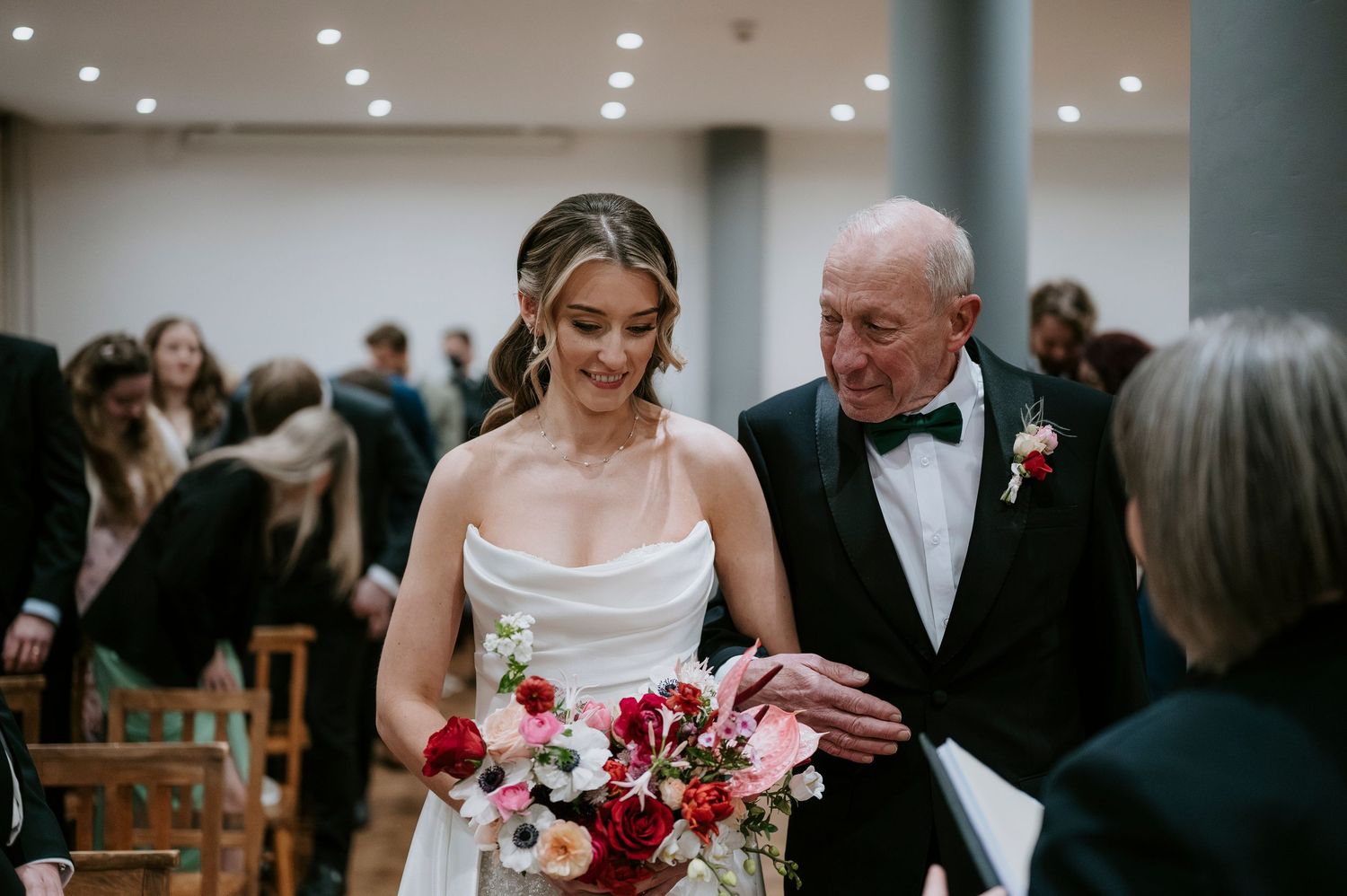 Wedding ceremony guests seated in rows of wooden chairs in an elegant indoor venue with white brick walls.