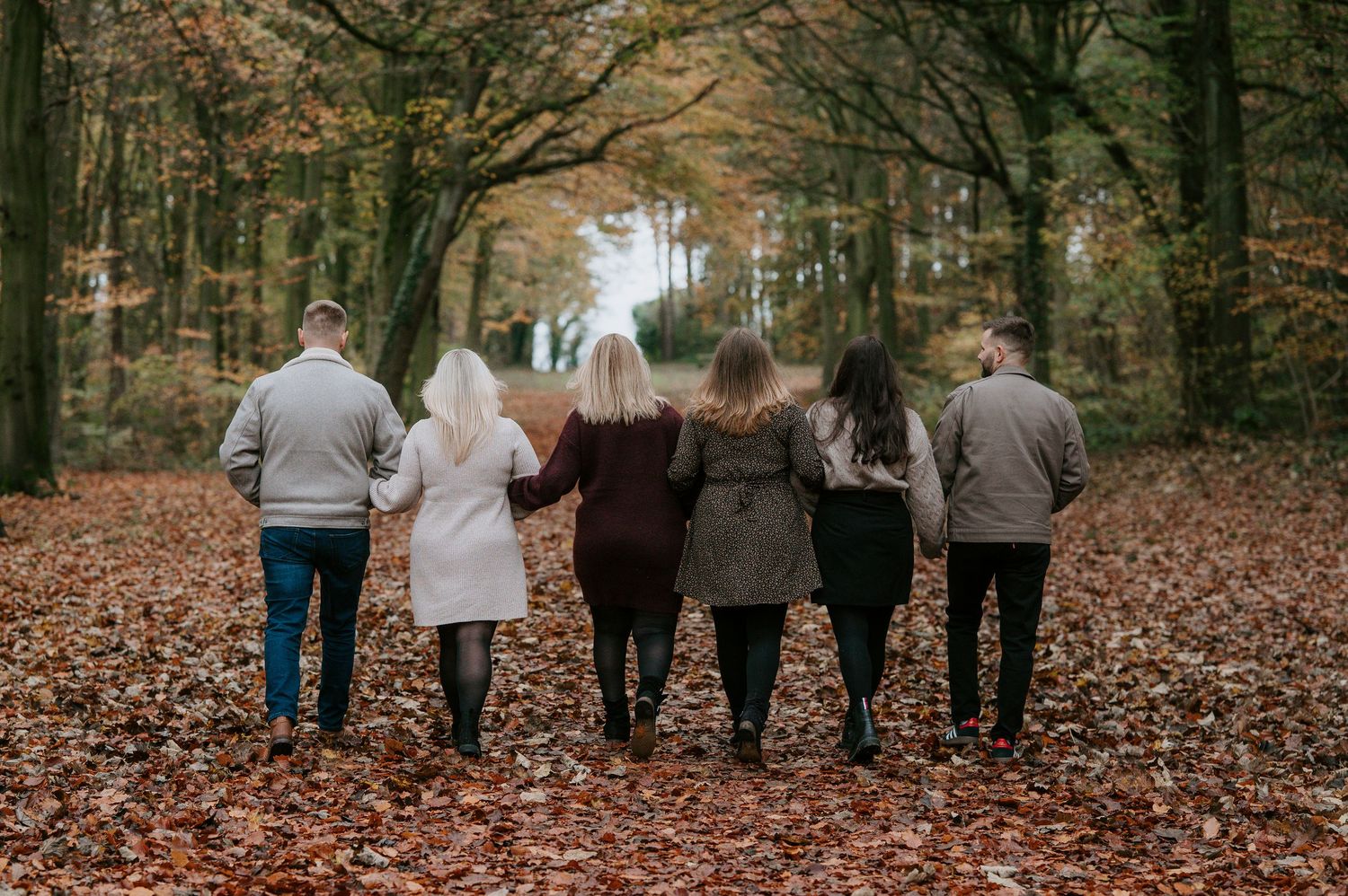 Group walking together arm in arm down a leaf-covered forest path during fall season.