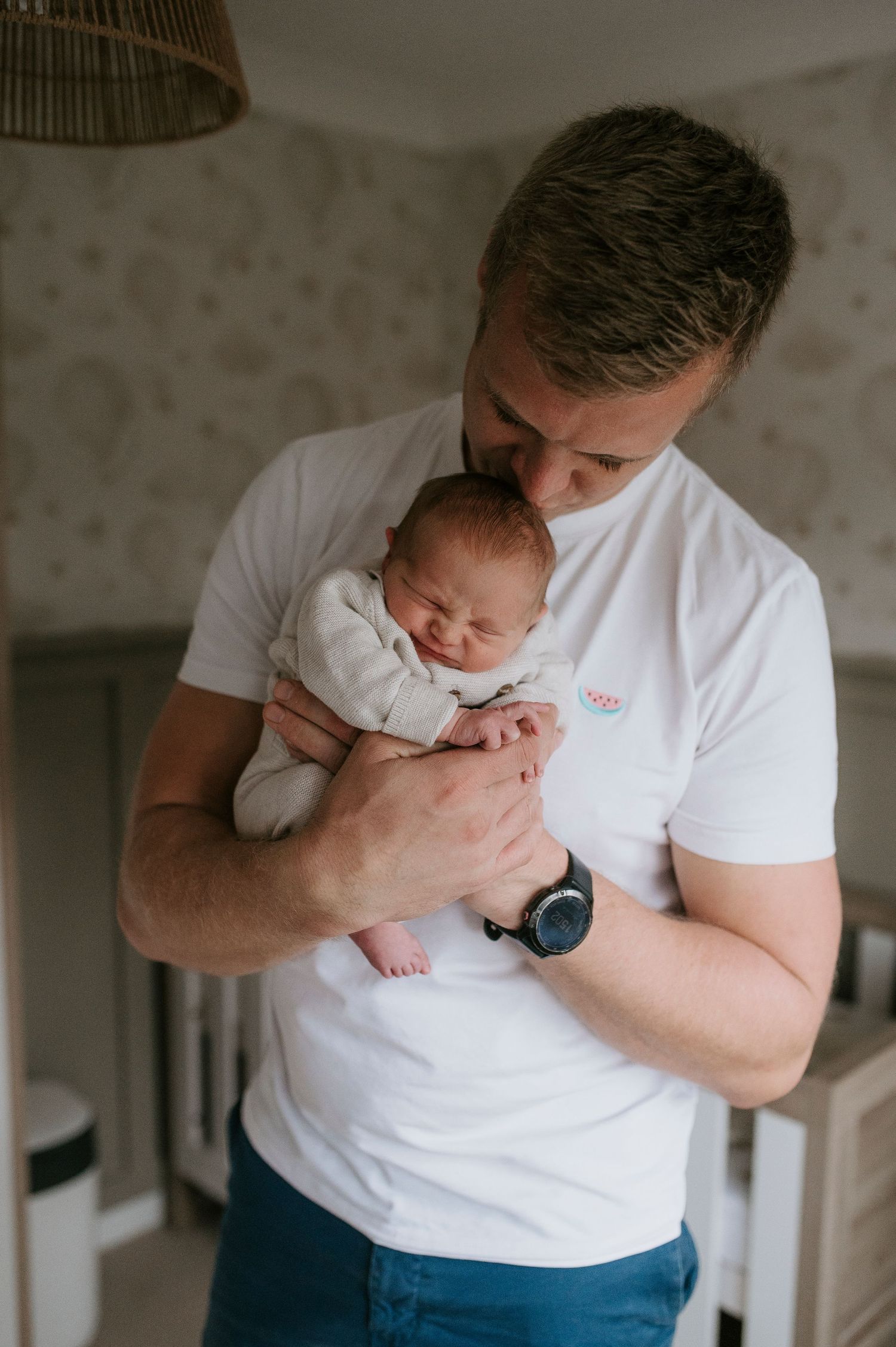 A person in a white t-shirt and jeans holds and kisses a baby in a series of tender indoor portrait shots.