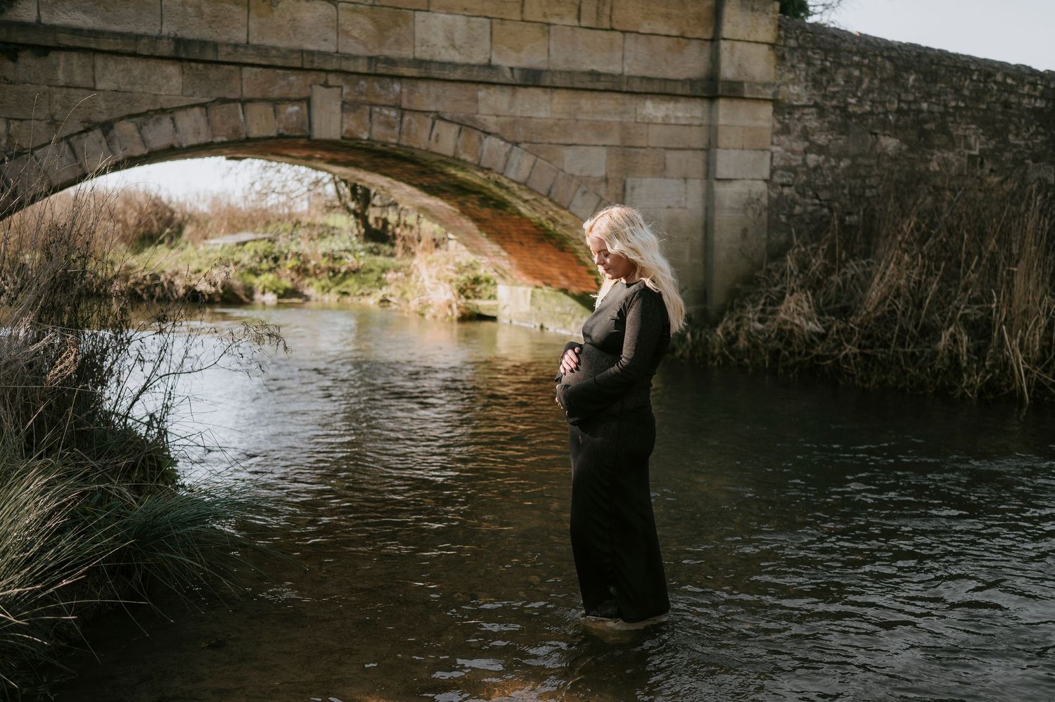 Person wading through shallow water under a stone bridge to explore the riverbed and arches.