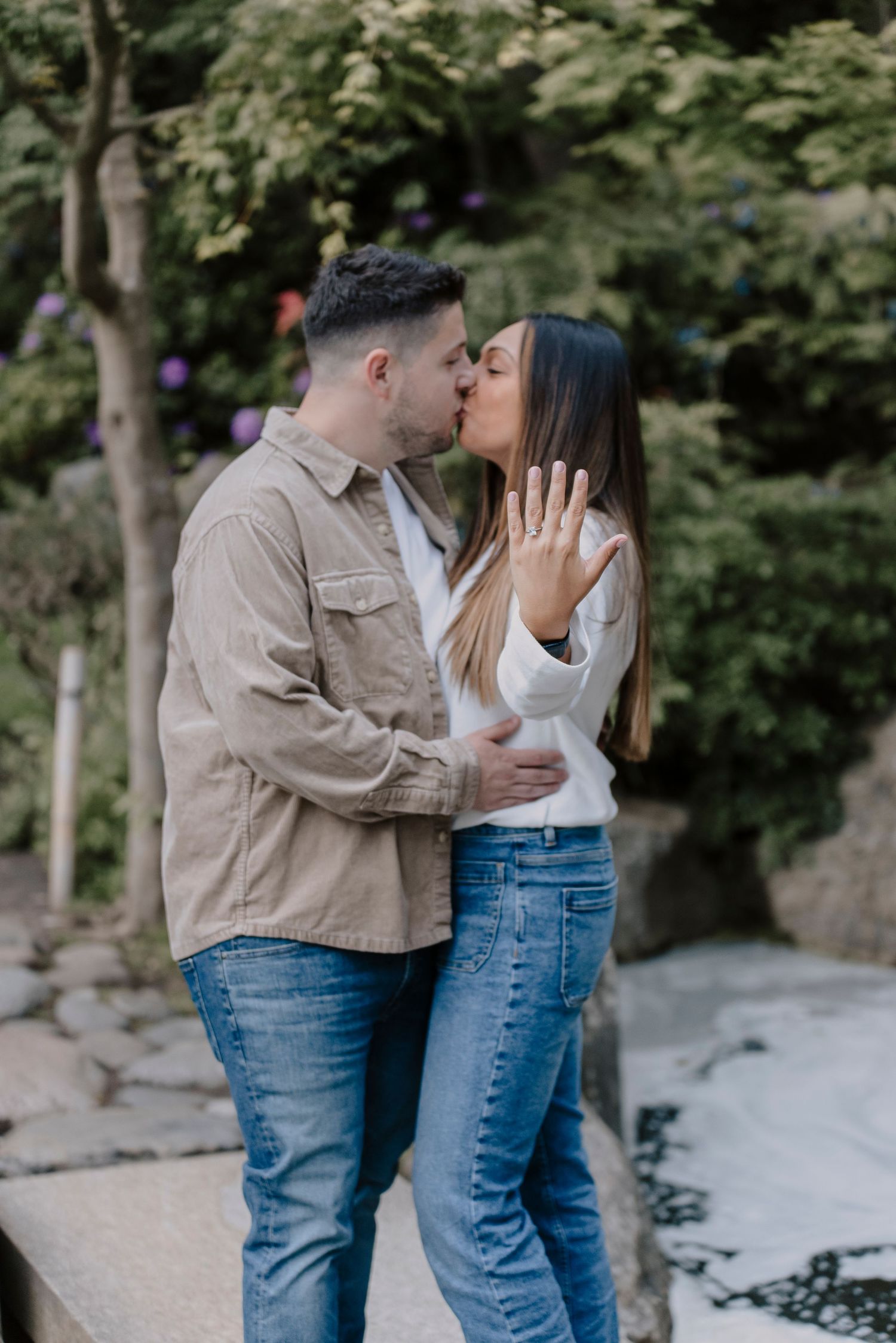 A couple embraces tenderly while standing on a stone path surrounded by lush greenery.