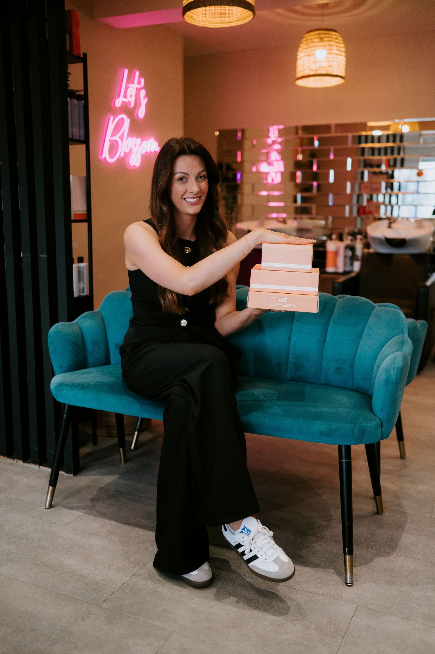 Woman in black dress sits on teal velvet sofa in modern room with pink neon lighting and pendant lamps.