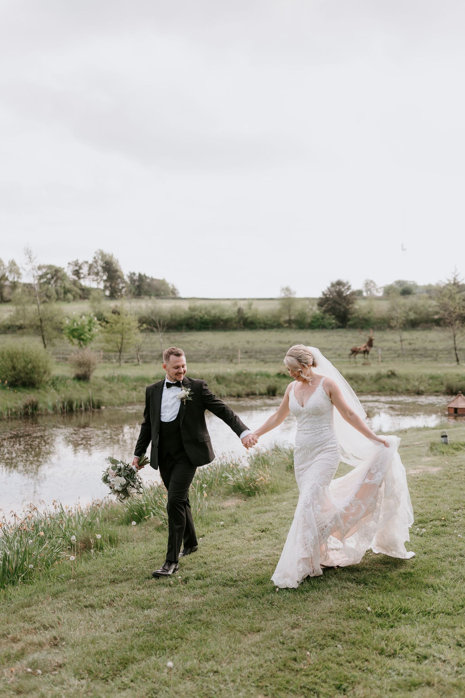 A couple in wedding attire runs playfully through a rustic field with a pond in the background.