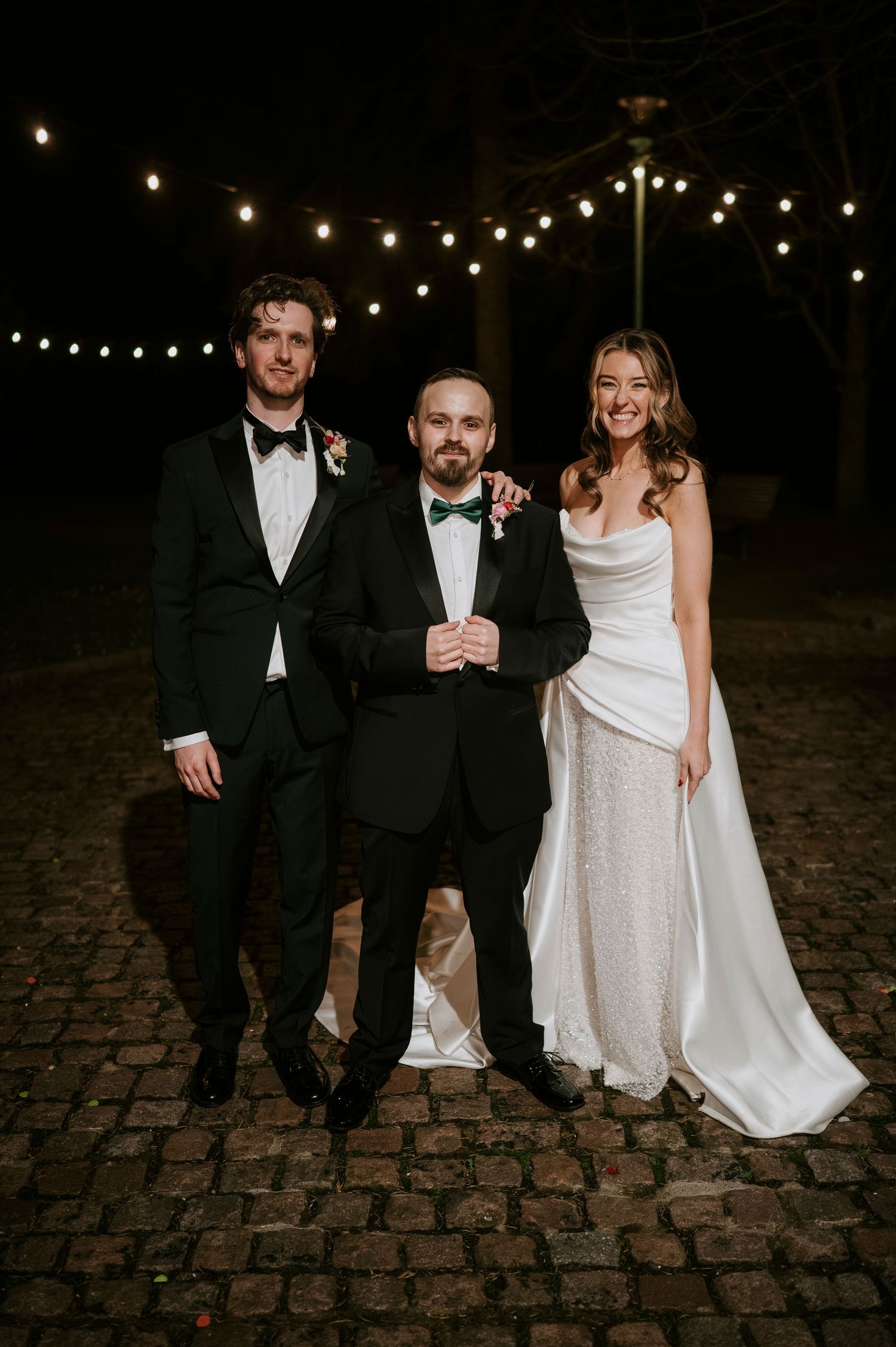 Wedding party members pose together on cobblestones under string lights during an evening celebration.