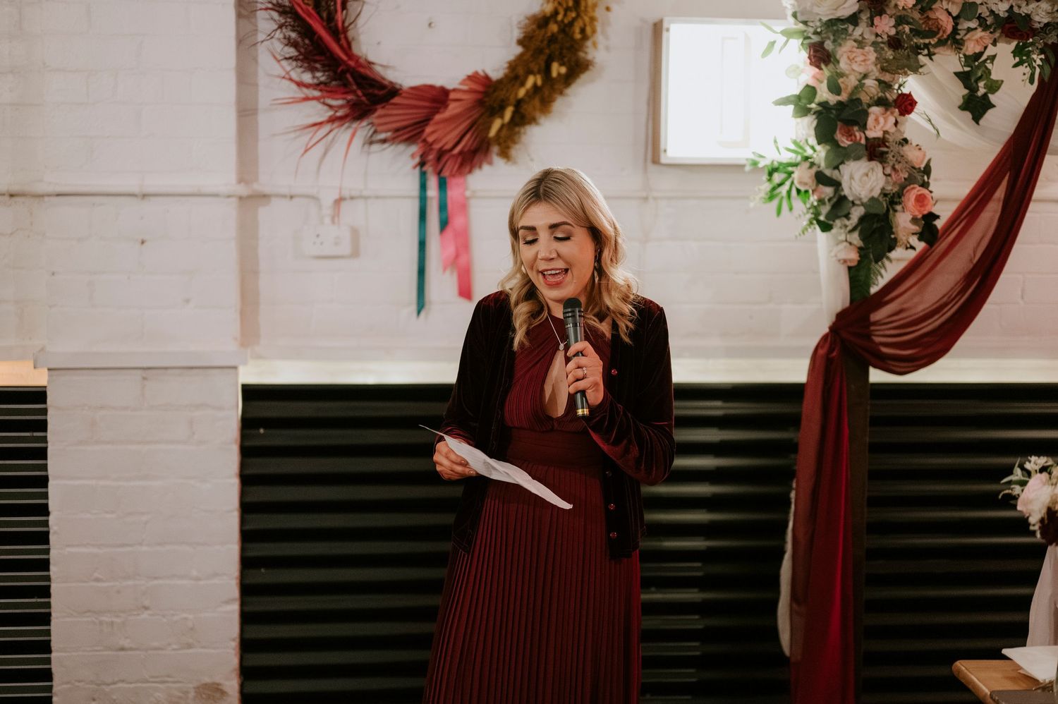 Blonde woman in burgundy pleated dress reads speech into microphone at wedding reception with floral decorations.