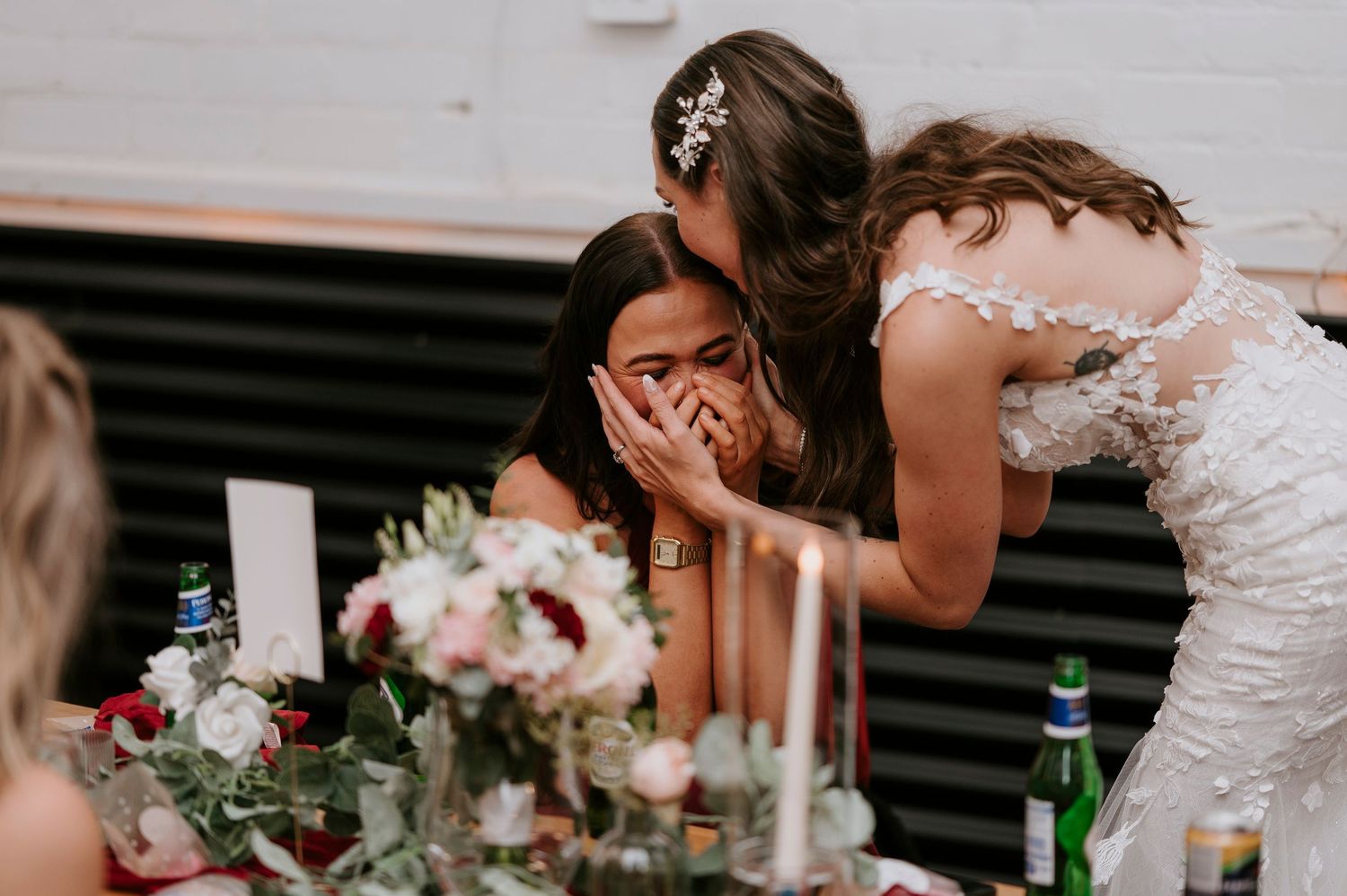 Emotional wedding guest covers her face while bride leans in to share a touching moment at the reception table.