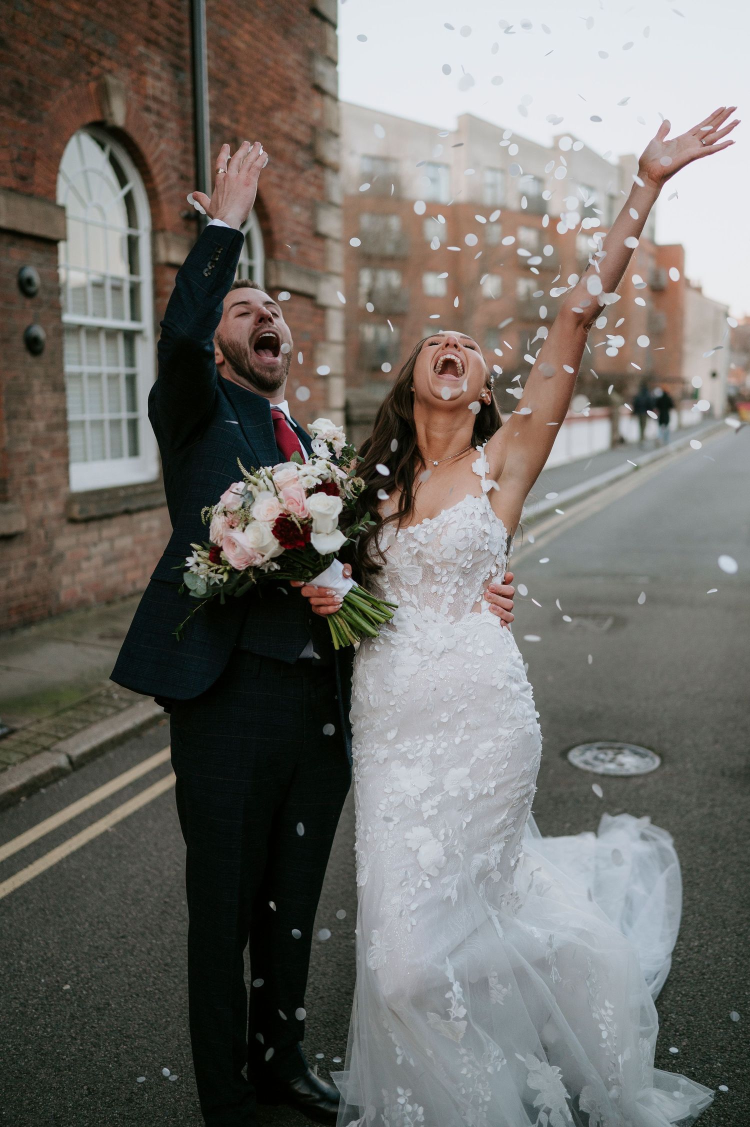 Joyful bride and groom celebrating with confetti toss on a street, bride in lace gown holding a bouquet.