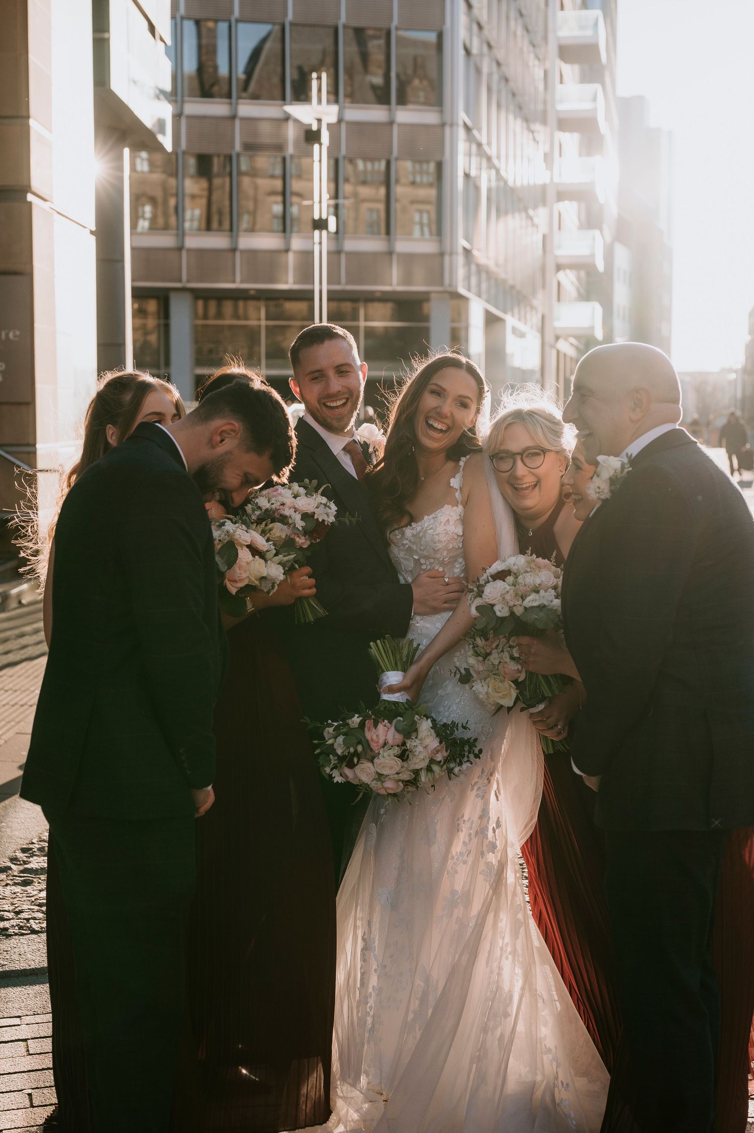 A joyful bride and groom celebrate with family outdoors in golden sunlight, surrounded by white floral bouquets in an urban setting.