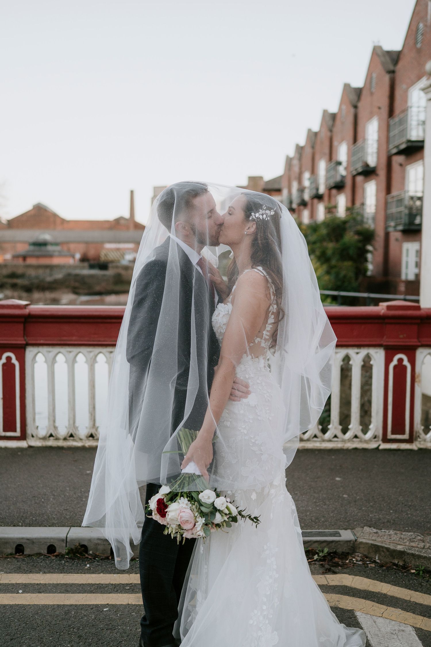 Bride and groom sharing a romantic kiss under a flowing veil on a bridge, bride holding a pink and red bouquet.
