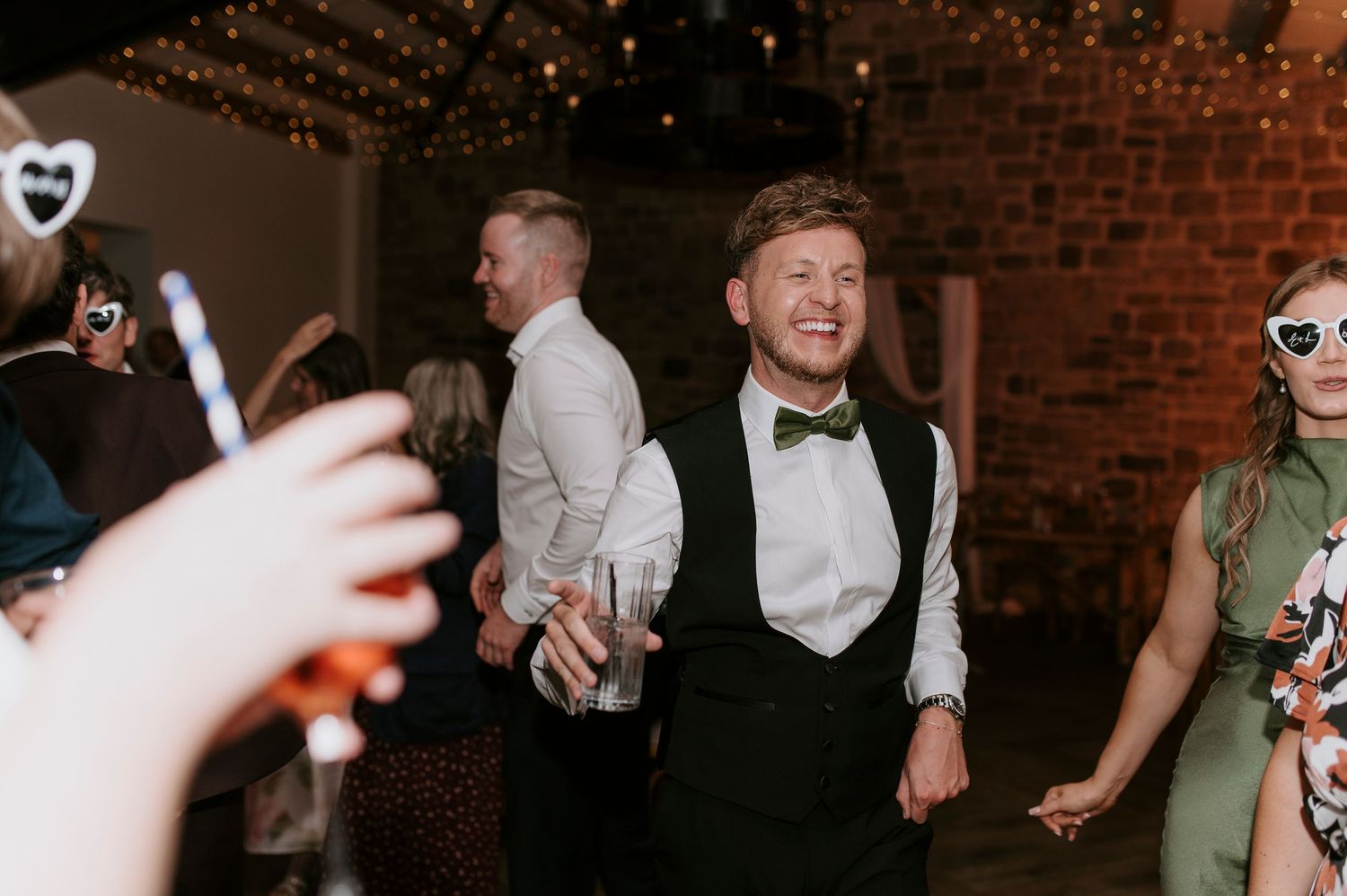 Wedding guests dance and celebrate on a dimly lit dance floor with exposed brick walls in the background.
