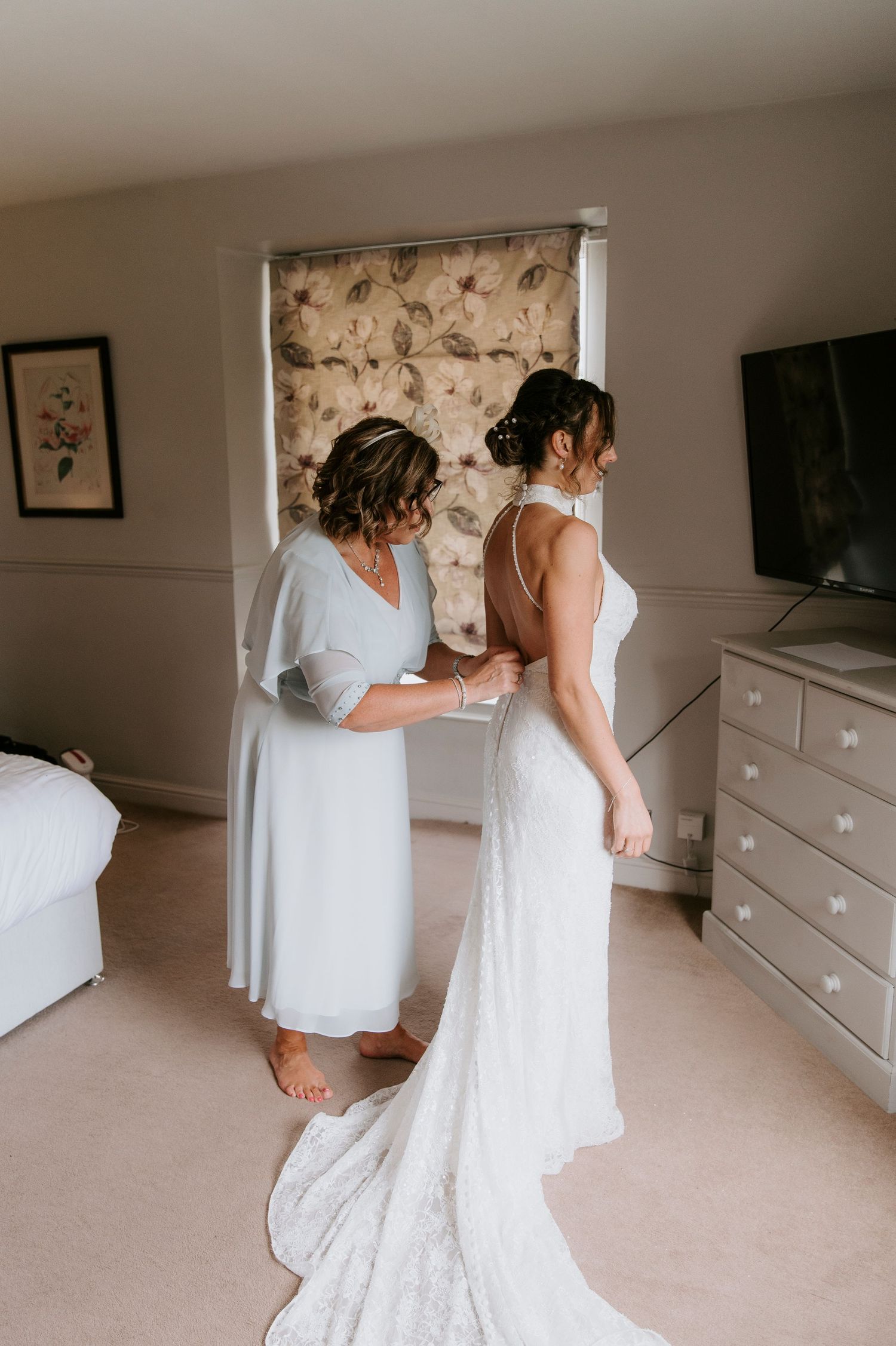 Two people in white dresses share an intimate moment while getting ready in a bedroom with light carpeting.