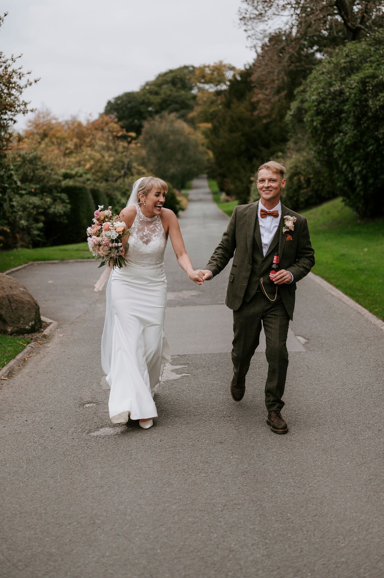 A couple in wedding attire walks playfully down a tree-lined path on their special day.