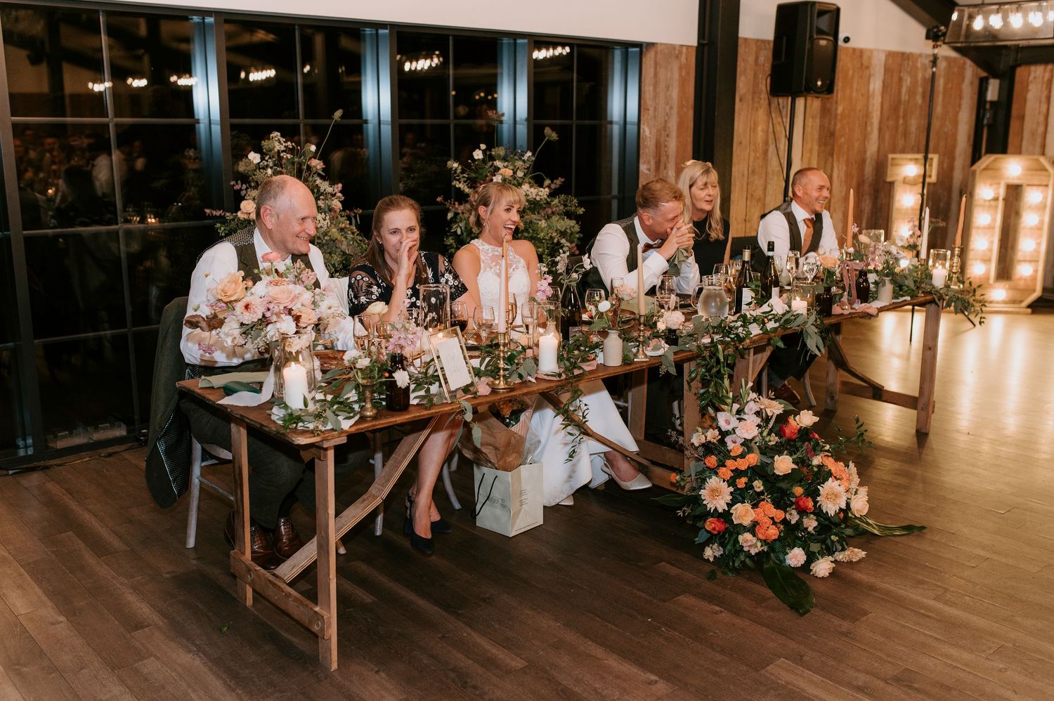 Wedding reception guests seated at long wooden tables in a rustic venue with warm lighting and floral centerpieces.