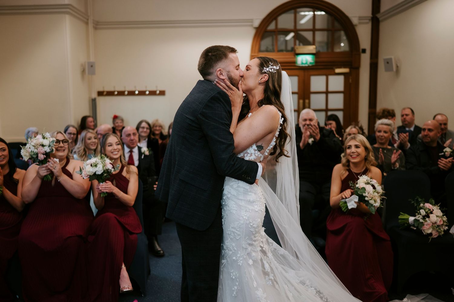 Bride and groom share first kiss at wedding ceremony while bridesmaids in burgundy dresses and guests applaud.