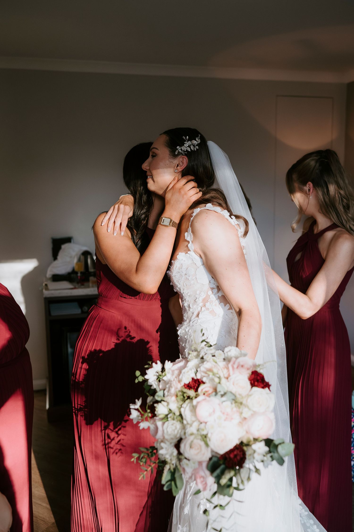 Bride in lace wedding dress embracing bridesmaid in burgundy gown while holding bouquet.
