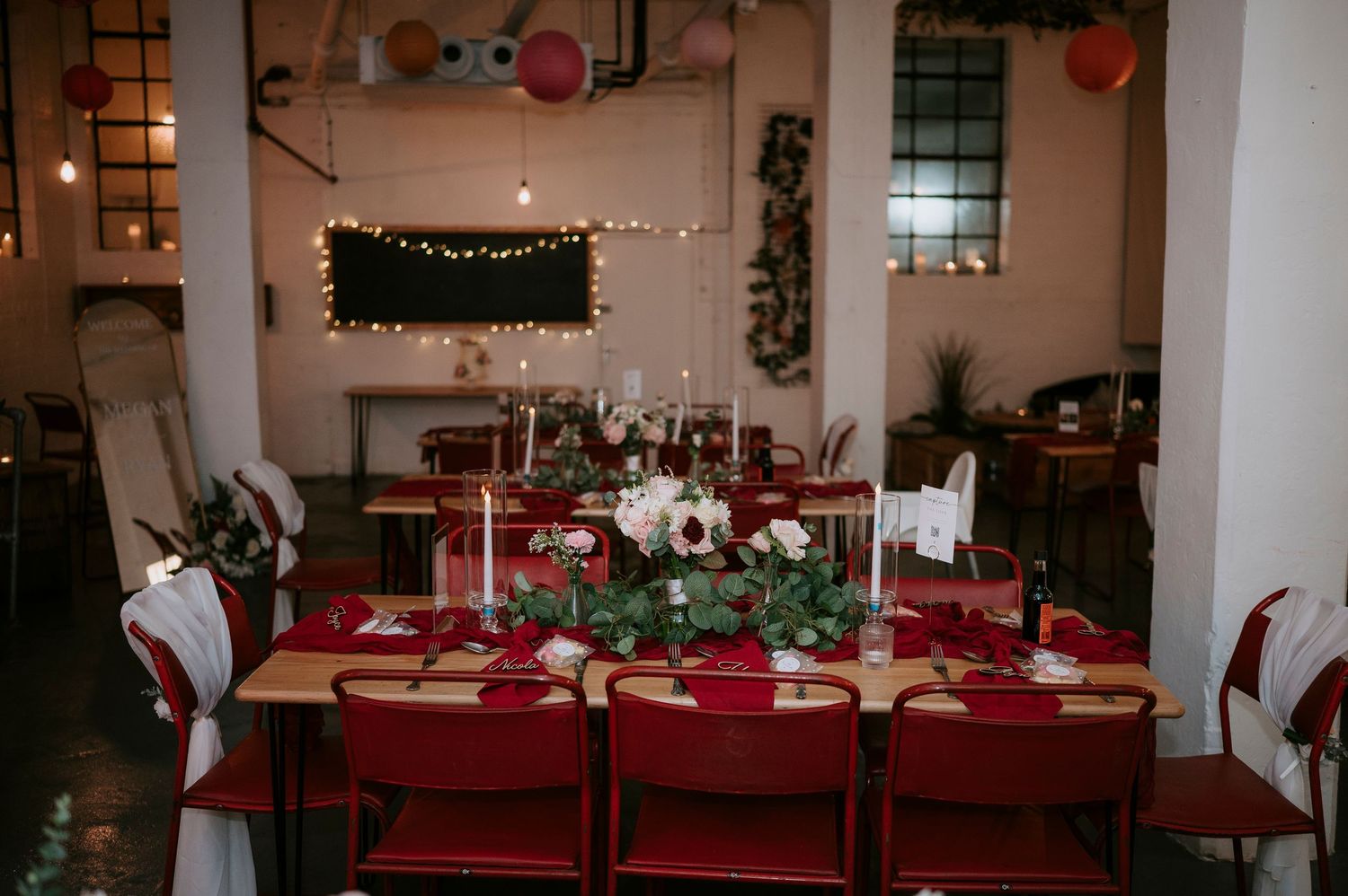 Elegant wedding reception table with red chairs, crimson napkins, candles, and lush greenery centerpiece in an industrial venue.