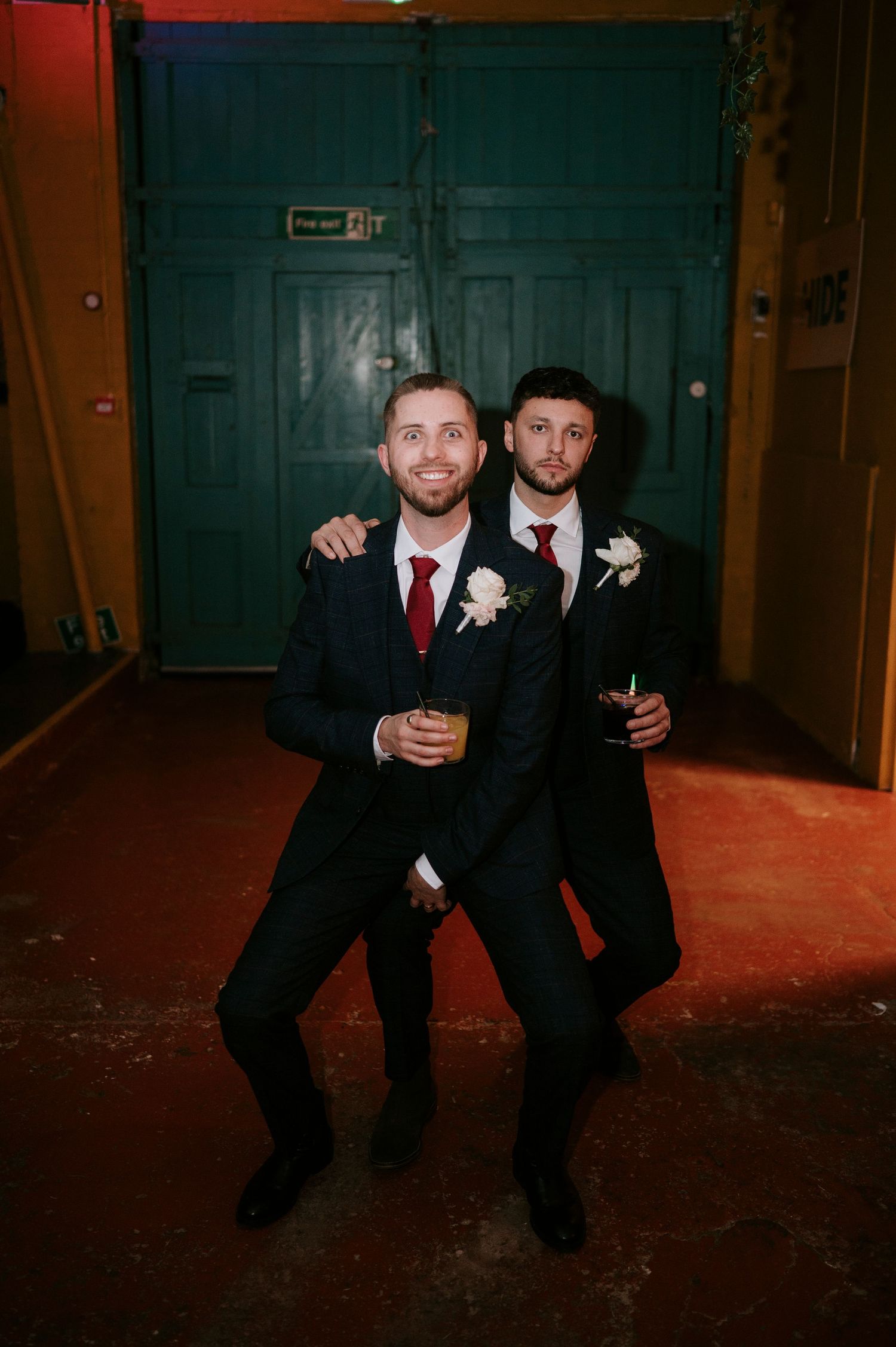 Two groomsmen in navy suits with red ties and boutonnieres pose playfully at a wedding reception.