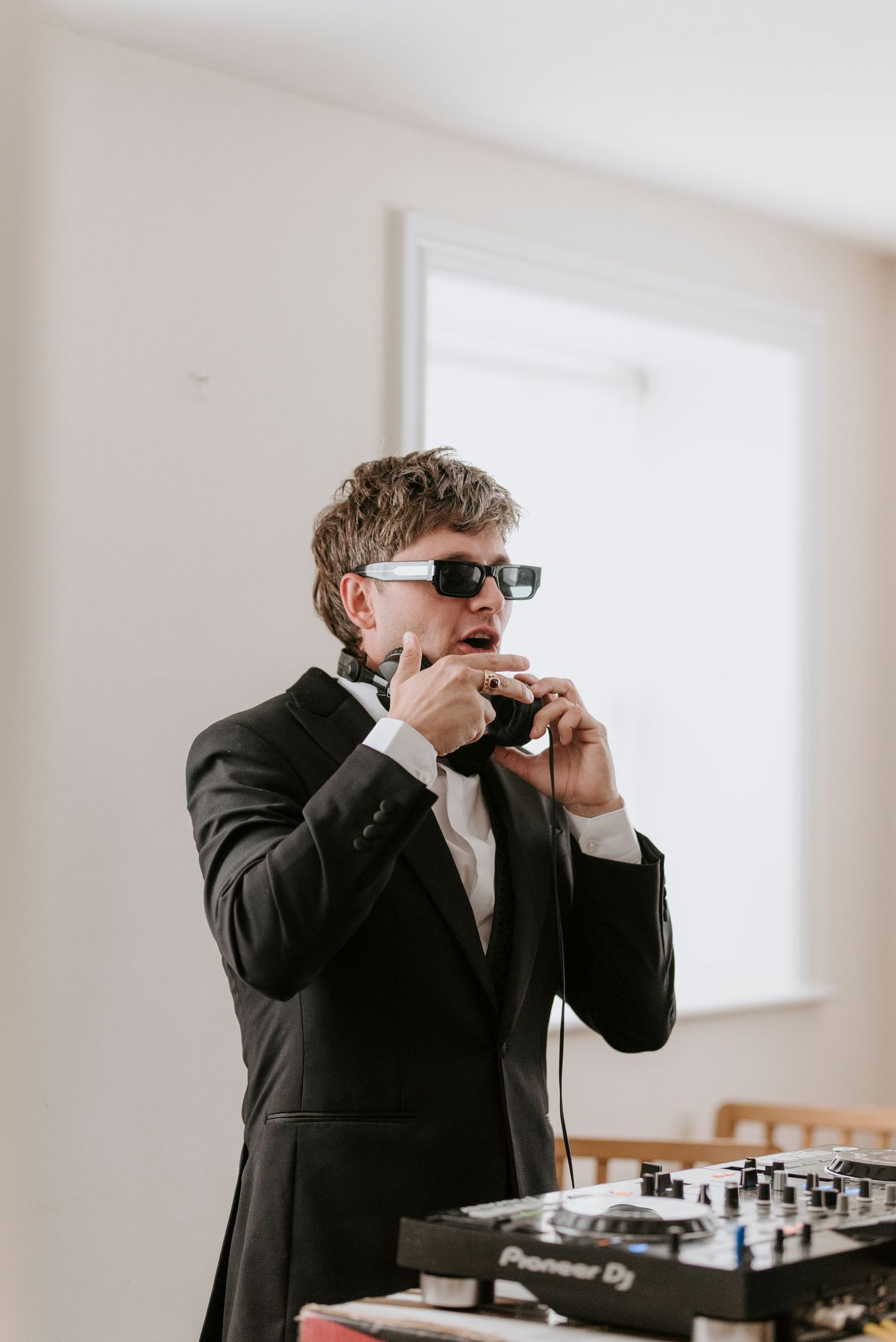 A DJ in a black suit and sunglasses stands at their mixing equipment during an event.